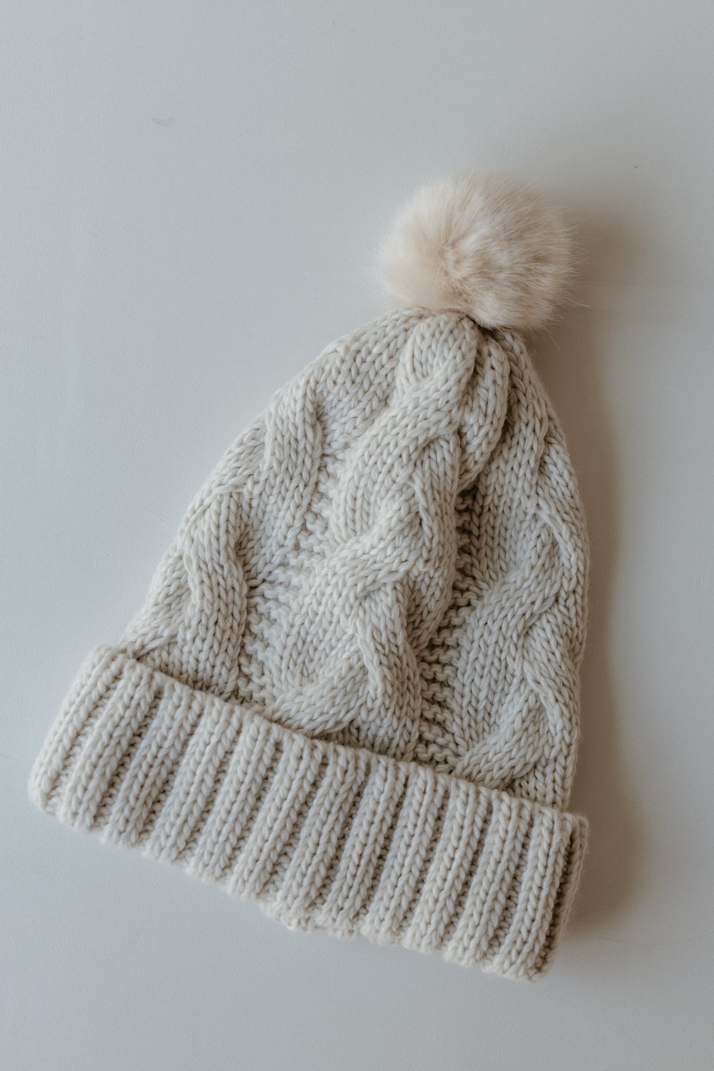 A cream-colored cable knit stocking hat with a folded brim and a fluffy pom-pom detail sits elegantly on a light surface, making it the perfect winter accessory.
