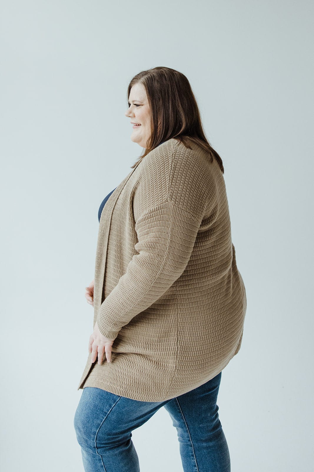 A person standing sideways, dressed in Yesta's Knit Stripe Pattern Open Cardigan in Light Taupe paired with blue jeans, against a plain light background.