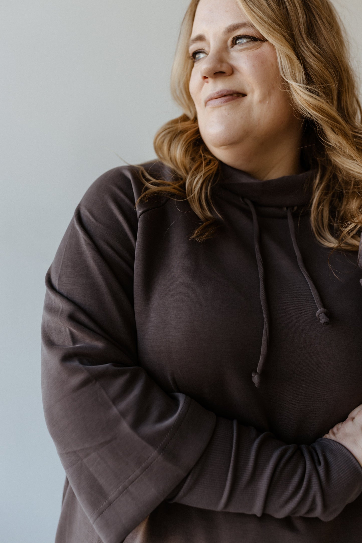 Person wearing a dark cowl neck tunic, smiling softly, and looking to the side against a plain background, exuding luxurious comfort.