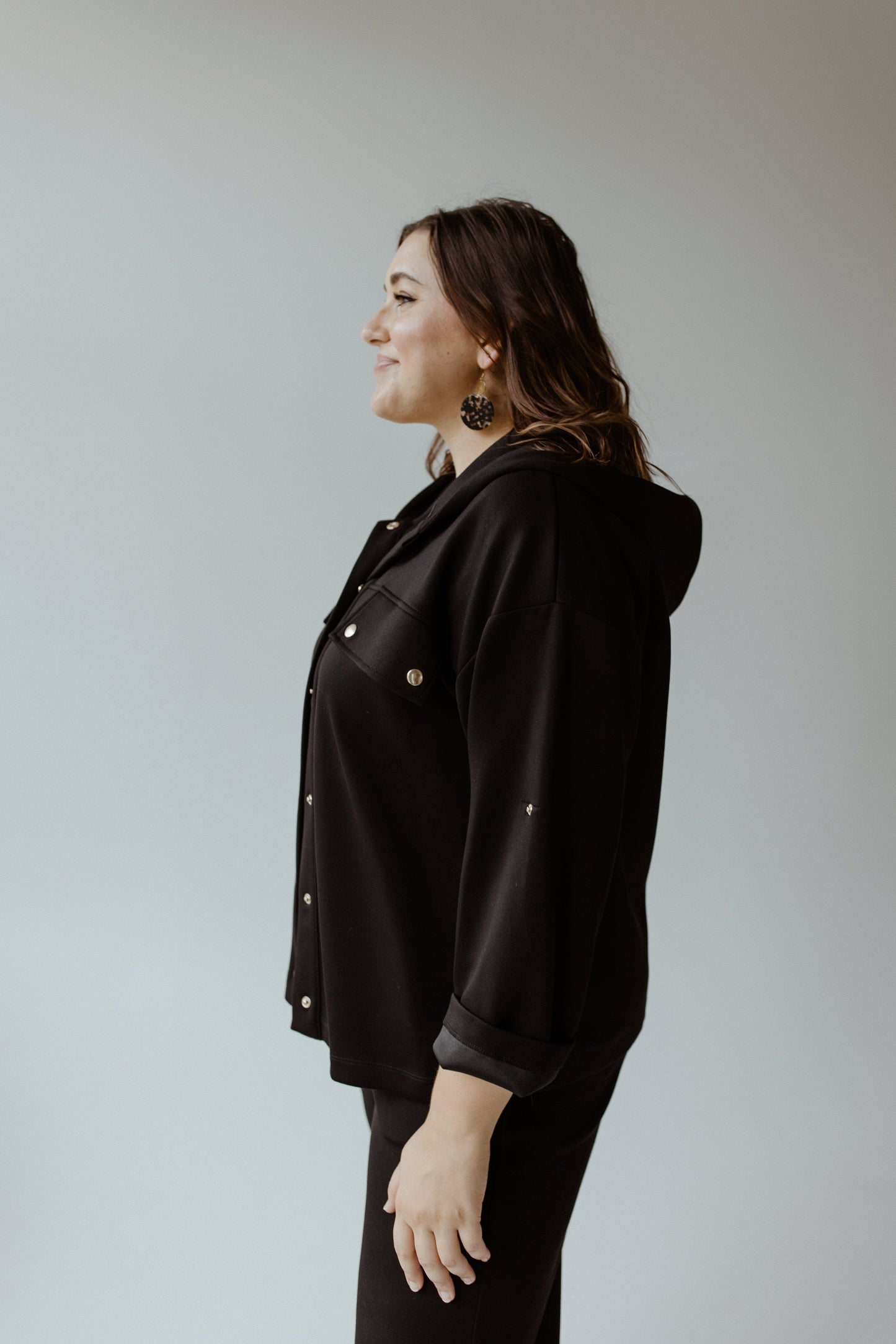 A person stands in profile, showcasing a sleek black jacket with button details—a perfect piece of contemporary leisurewear—against a light gray background.