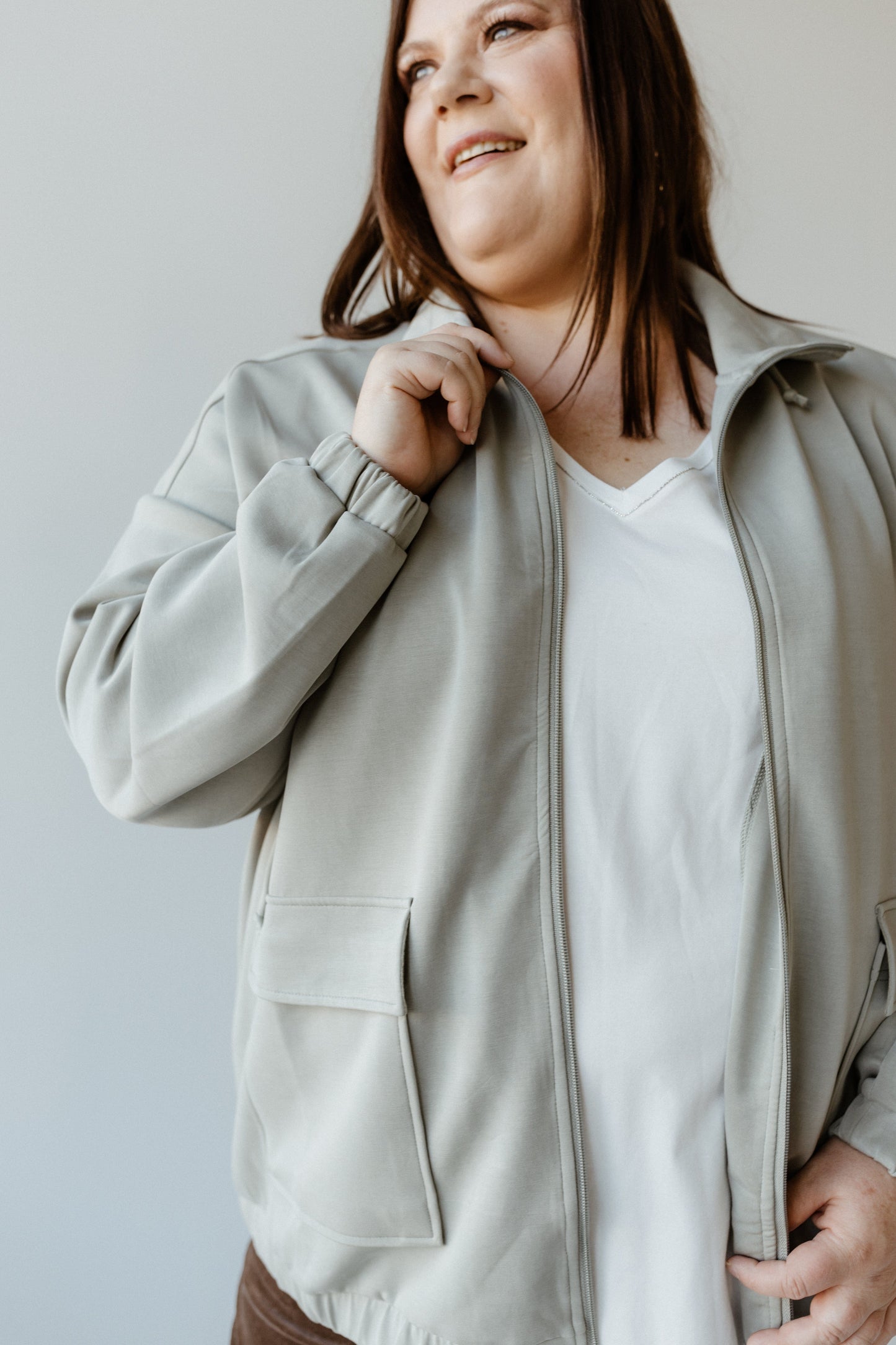 Wearing the LIGHT AS AIR ZIP-UP JACKET in an elegant Evergreen Fog shade, a person smiles warmly while looking away from the camera, effortlessly combining fashion and function.