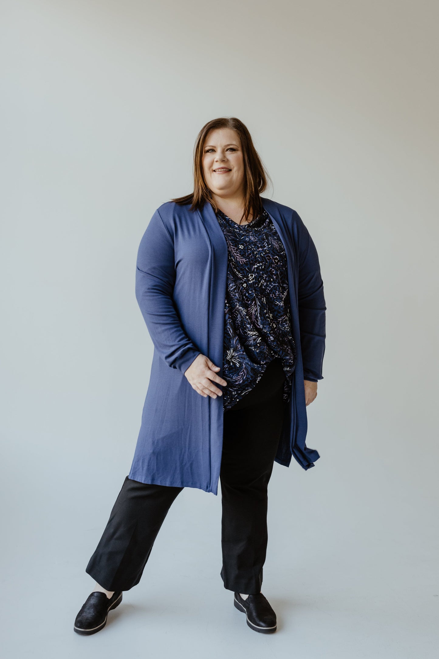 A person stands against a plain background wearing the Yesta LONG SLEEVE CARDIGAN WITH BACK RUCHING IN TURKISH SEA, paired with a patterned top, black pants, and black shoes.