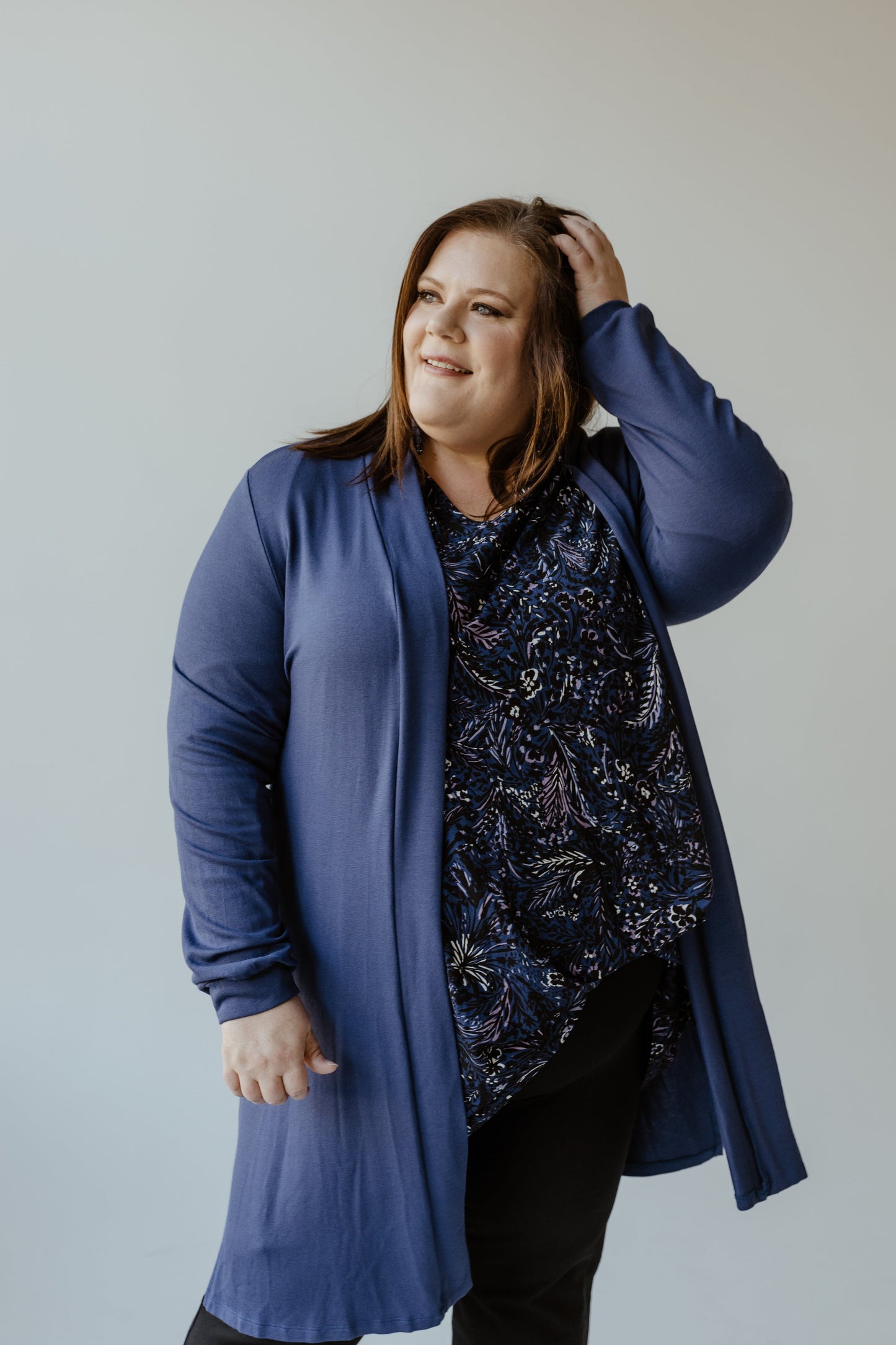 A person poses against a neutral backdrop, donning the Yesta Long Sleeve Cardigan with Back Ruching in Turkish Sea over a patterned top, thoughtfully placing one hand on their head.