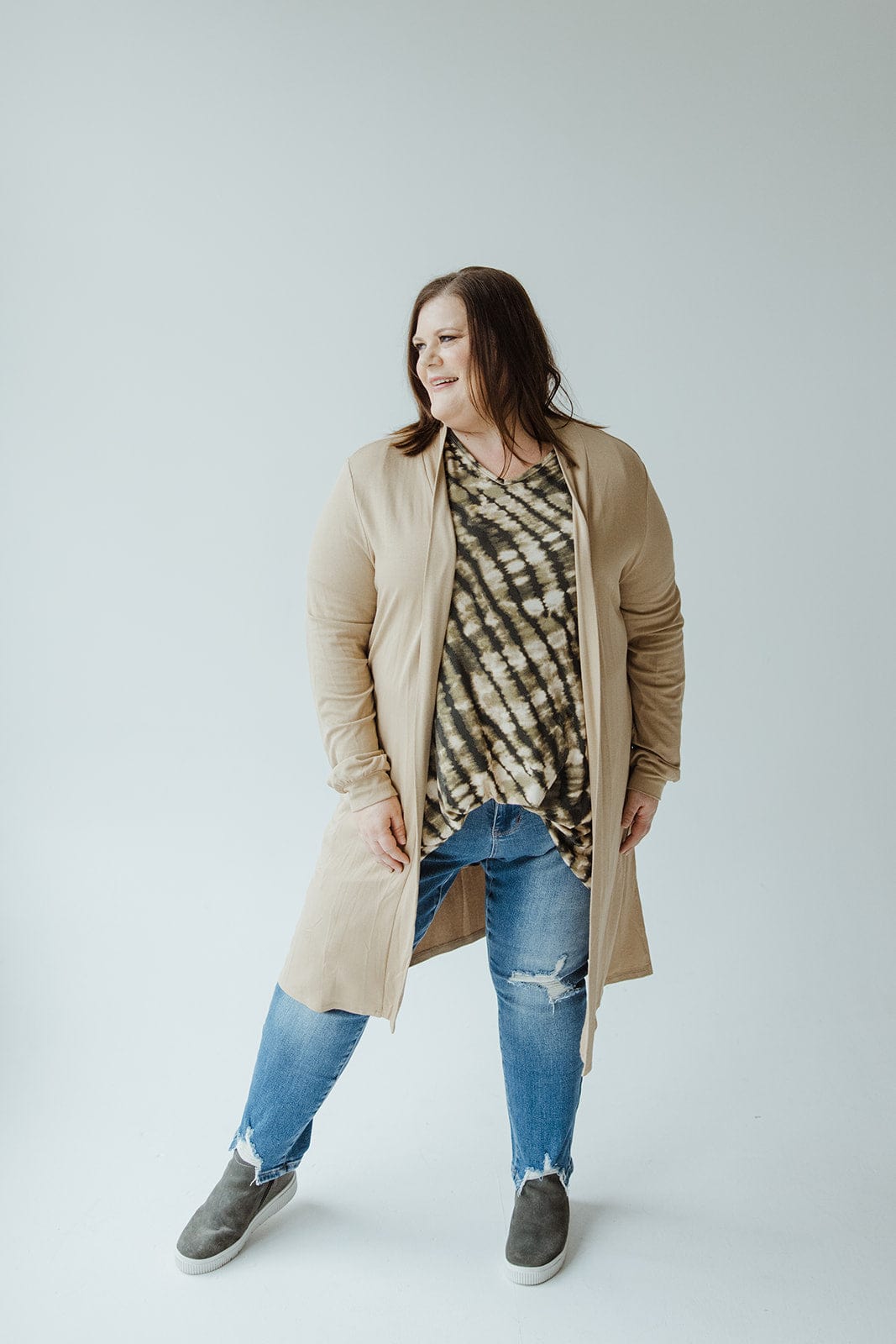 A person stands against a plain background, wearing the Yesta LONG SLEEVE CARDIGAN WITH BACK RUCHING IN WARM ALMOND over a patterned shirt, blue jeans with rips, and gray slip-on shoes.