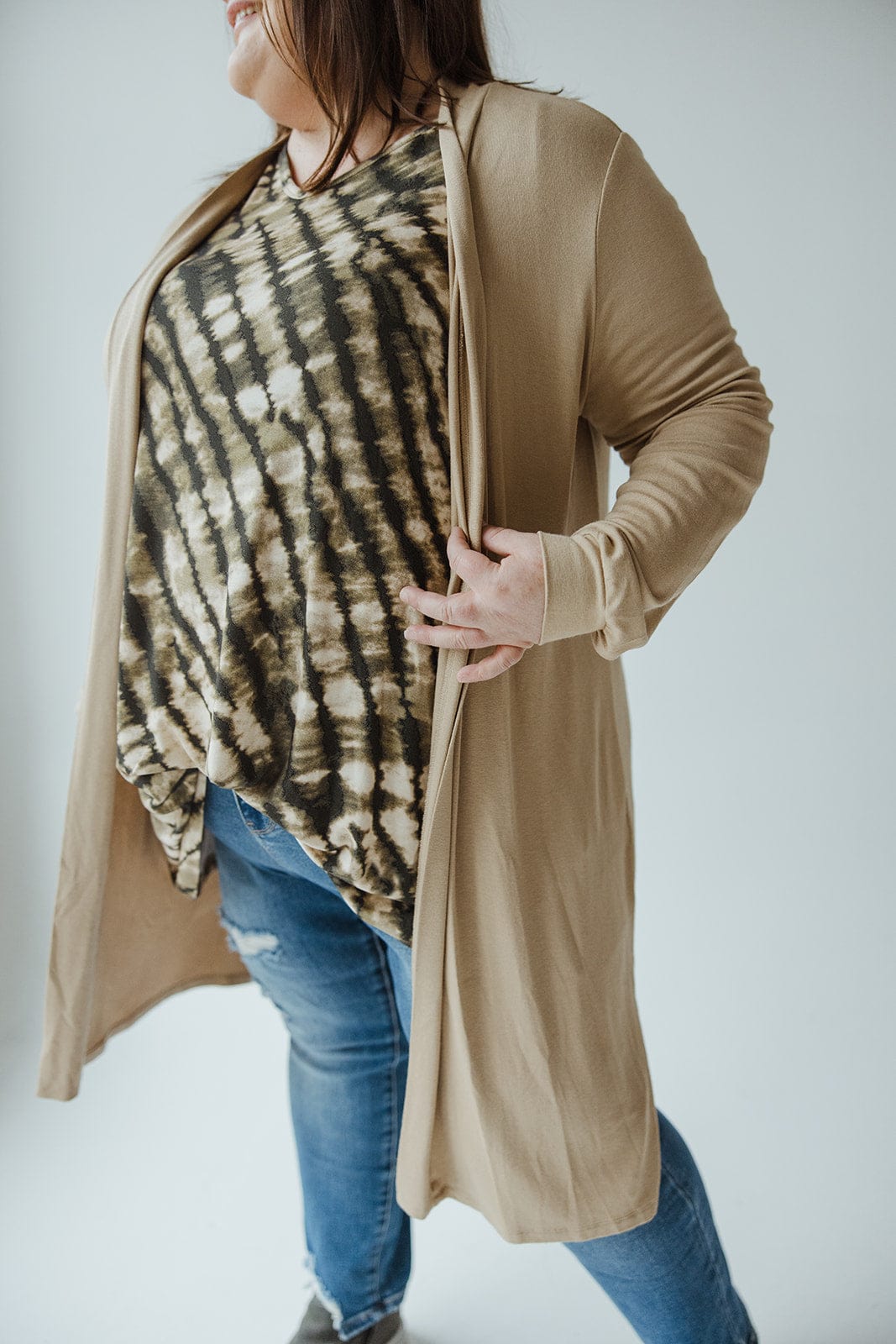 A person wearing the Yesta Long Sleeve Cardigan with Back Ruching in Warm Almond over a tie-dye shirt and blue jeans poses against a light background.