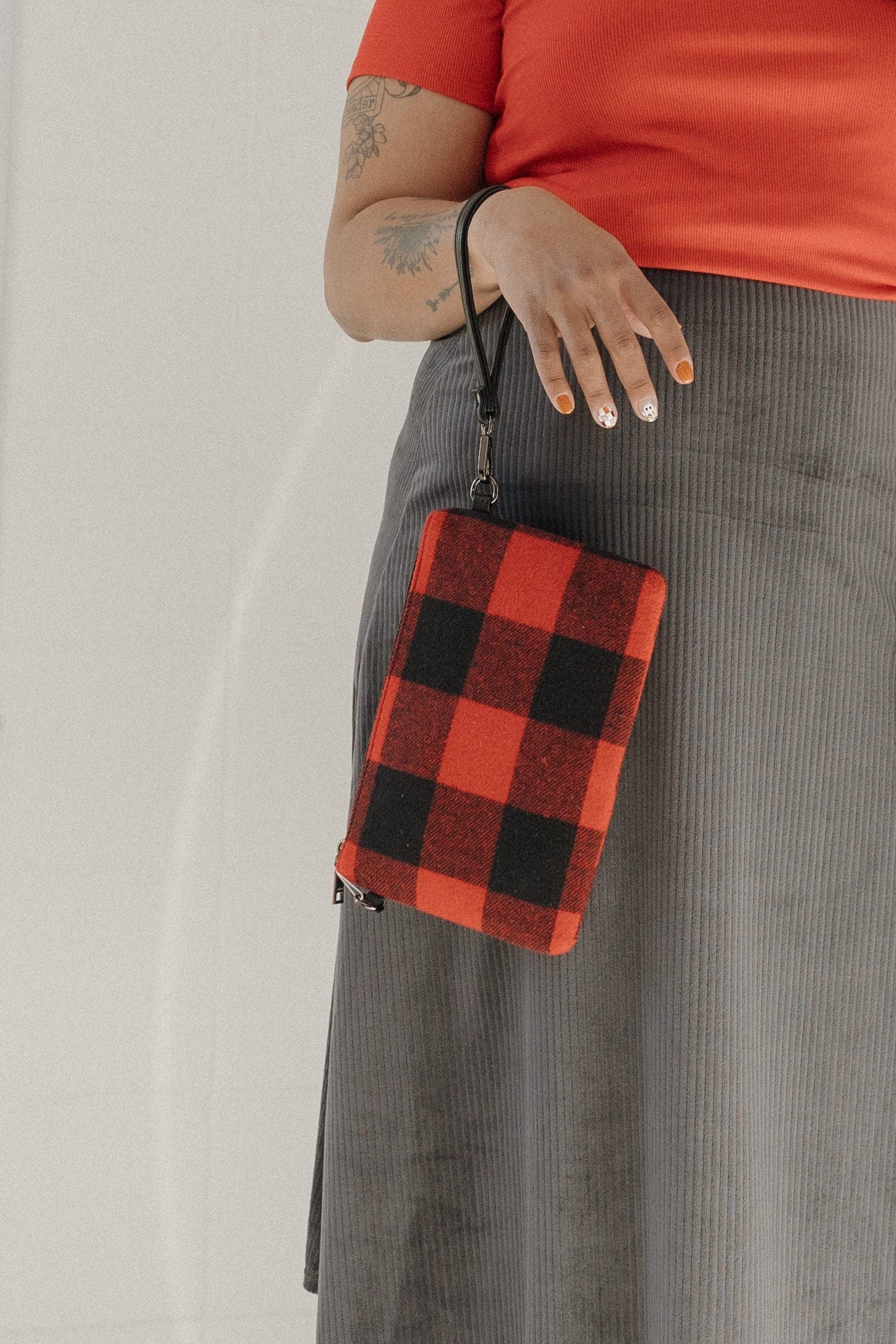 Person wearing a red shirt, gray skirt, and black boots holding a red and black checkered bag.