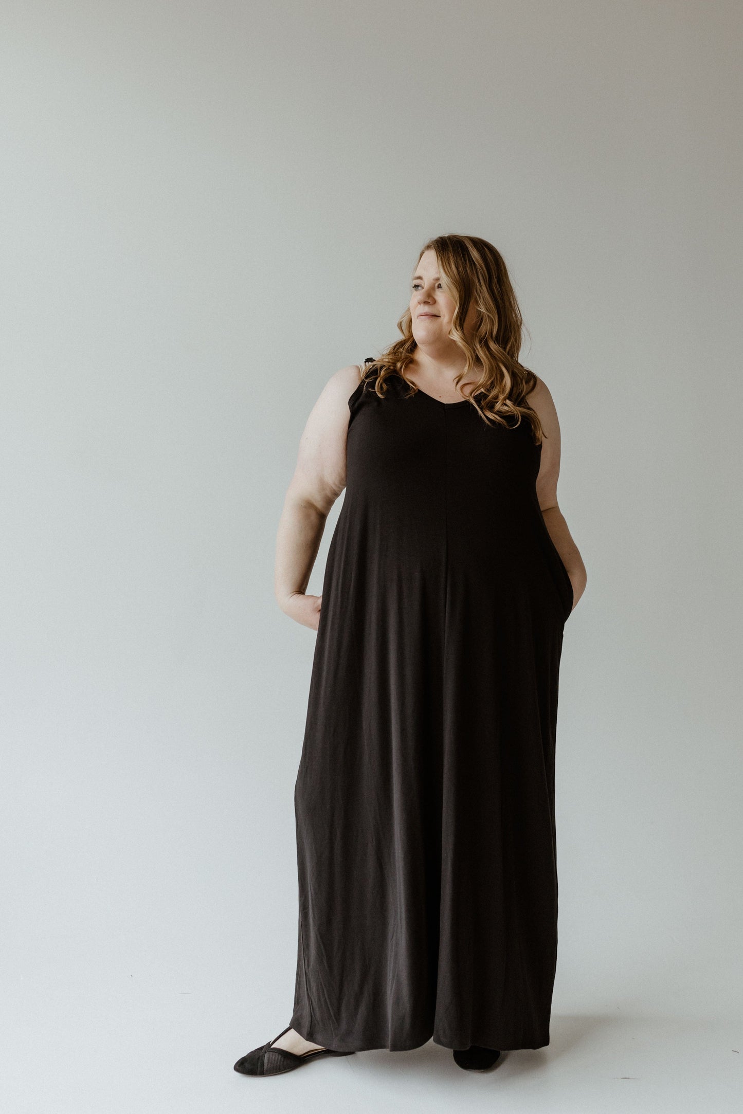 A person with long hair wearing a black sleeveless slip dress stands against a plain background, looking to the side with hands in pockets. The dress features adjustable cami straps, offering both style and comfort.