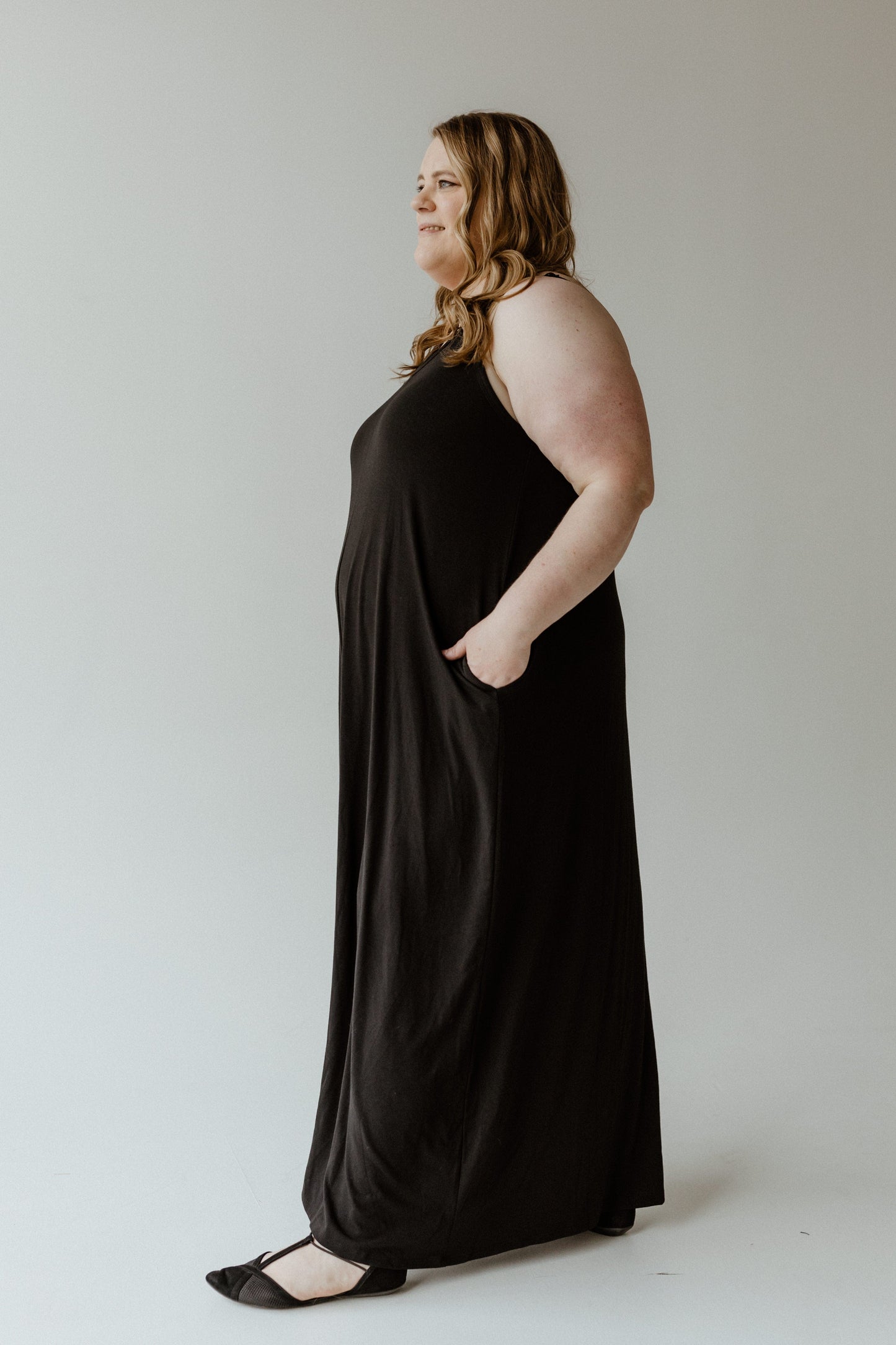 A woman in a long black slip dress stands in profile against a plain background, with her left hand in a pocket and her right hand relaxed. The adjustable cami straps add versatility to her elegant look, making this piece an essential part of any plus-size wardrobe.