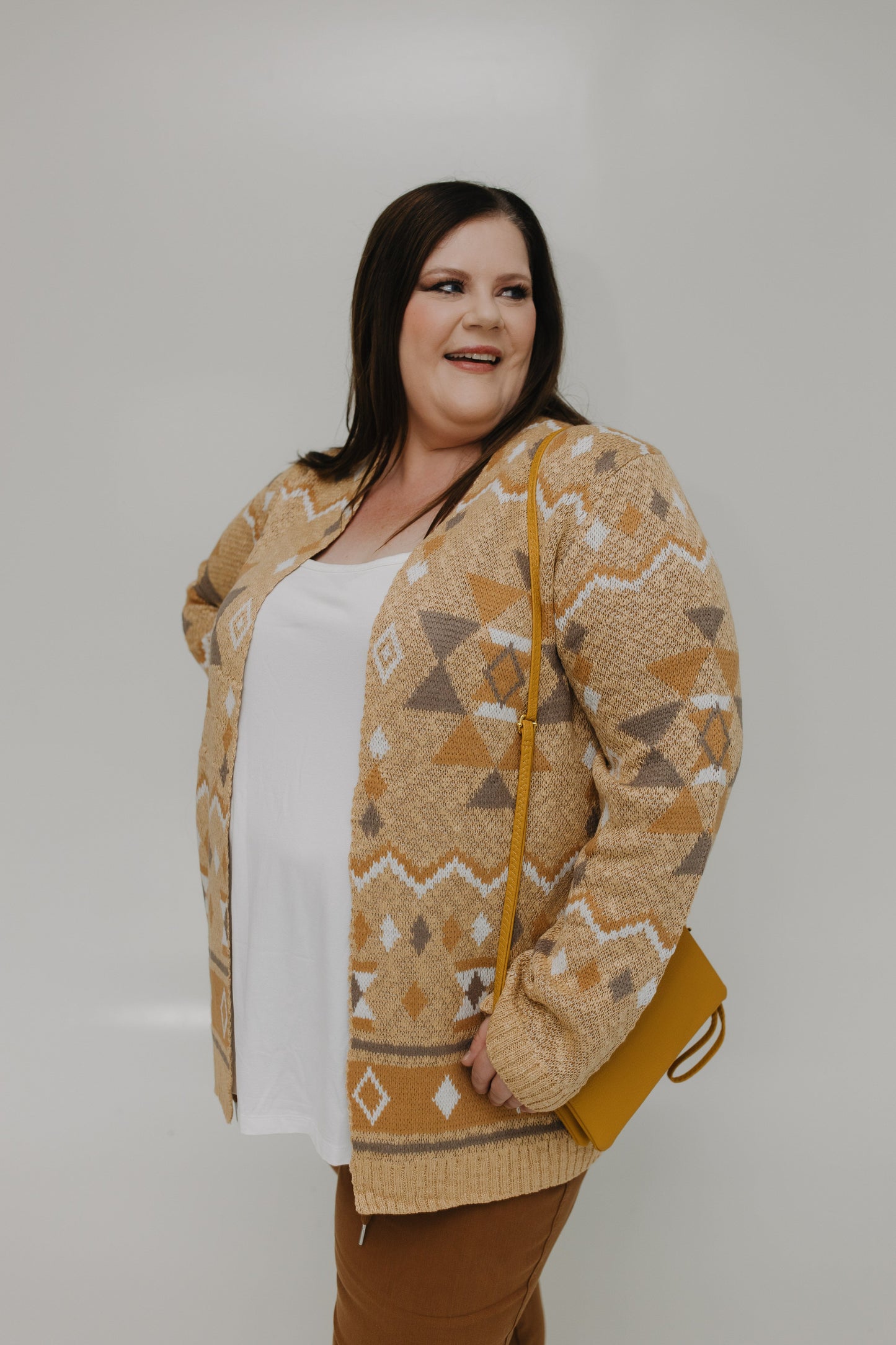 Woman wearing a patterned beige cardigan over a white top and brown pants, holding a mustard clutch against a plain background.