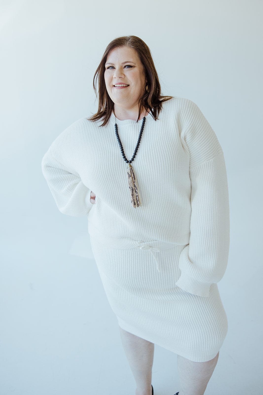 A person wearing a white sweater dress with subtle blouson sleeves and a long necklace stands against a light background, smiling warmly.