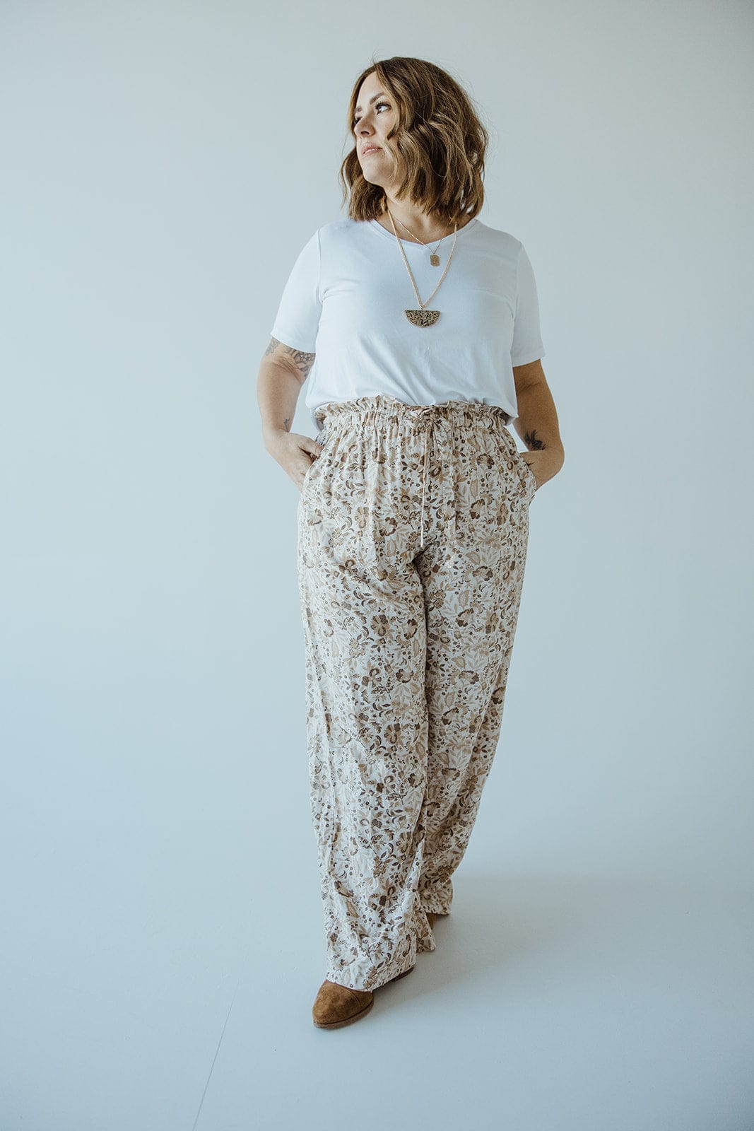 A woman stands against a plain background, wearing a white T-shirt, brown shoes, and Love Marlow's PAPERBAG WAIST WIDE LEG PRINTED PANTS. She has her hands in her pockets and is looking away from the camera.