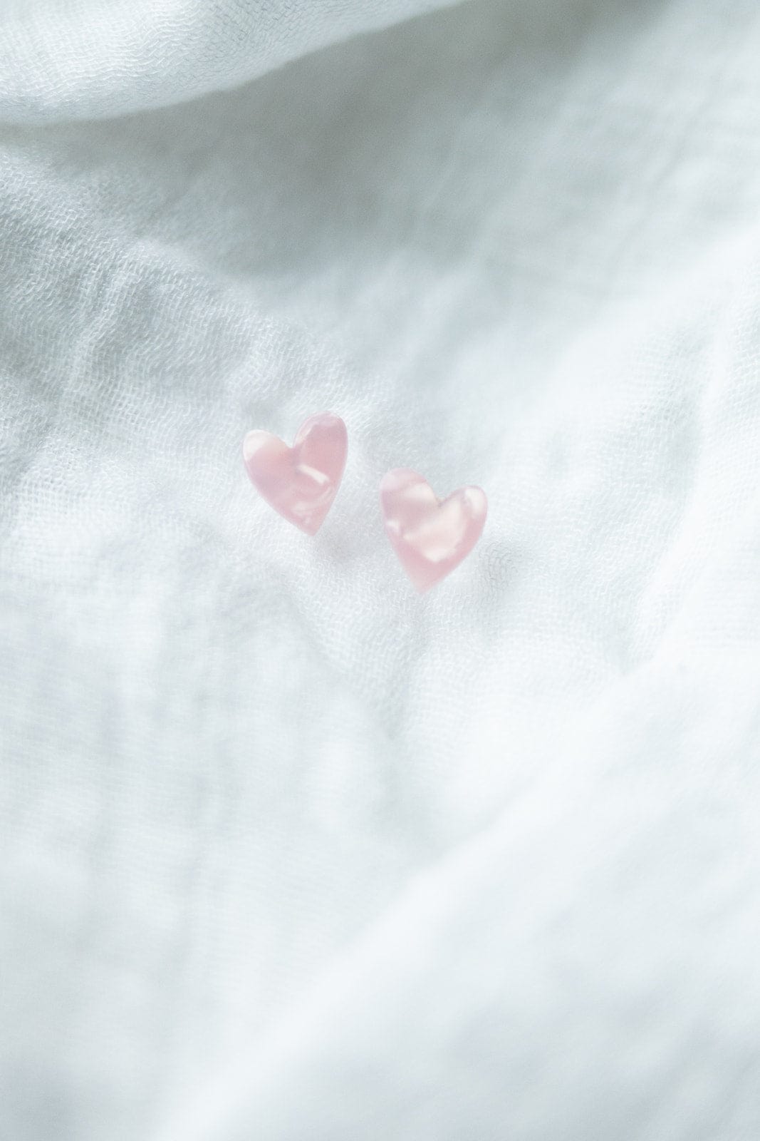 Two small pink heart studs rest elegantly on a textured white fabric background, crafted from plant-based acetate acrylic with hypoallergenic stainless steel posts for comfort and style.