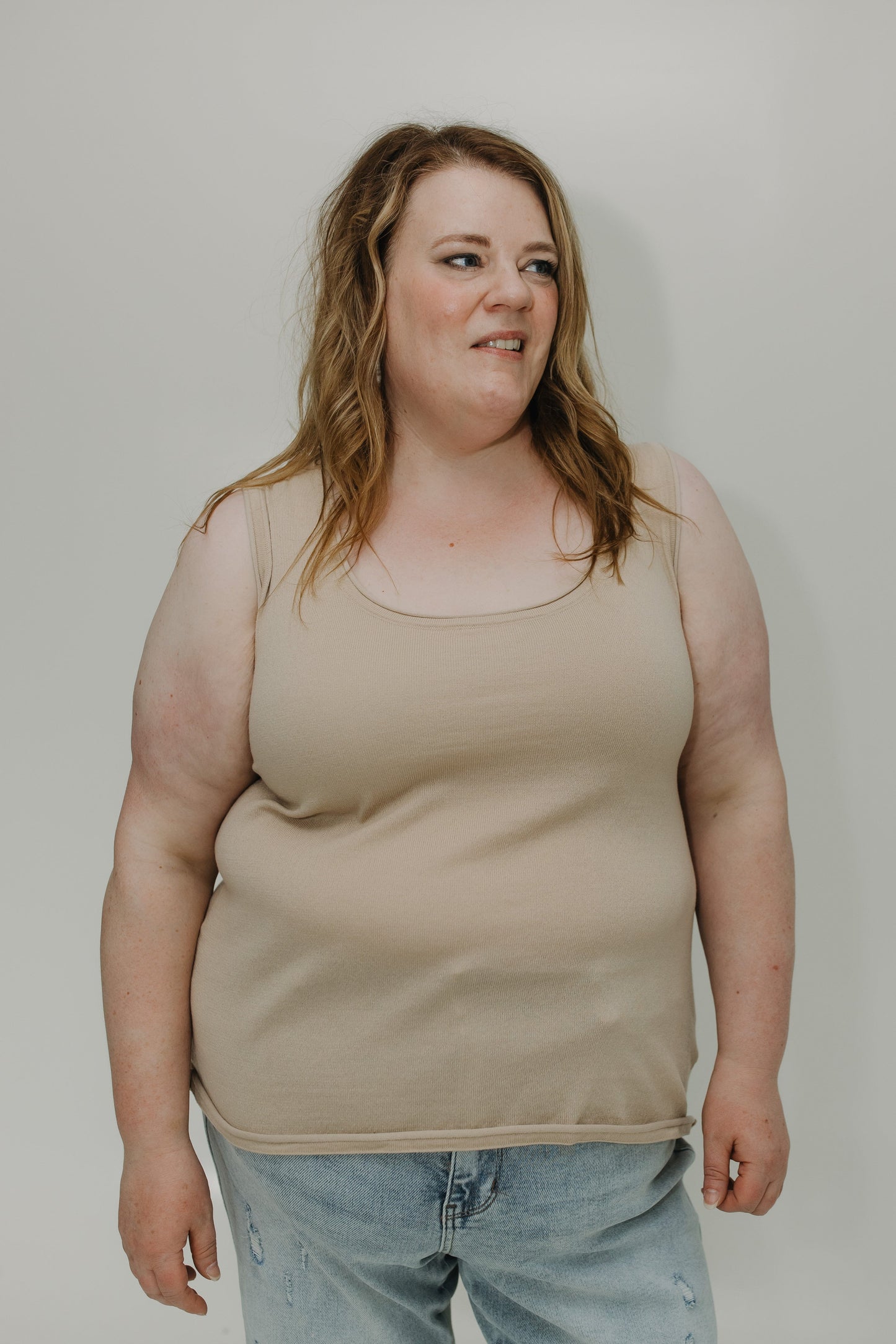 Woman wearing a beige tank top and light blue jeans against a plain background