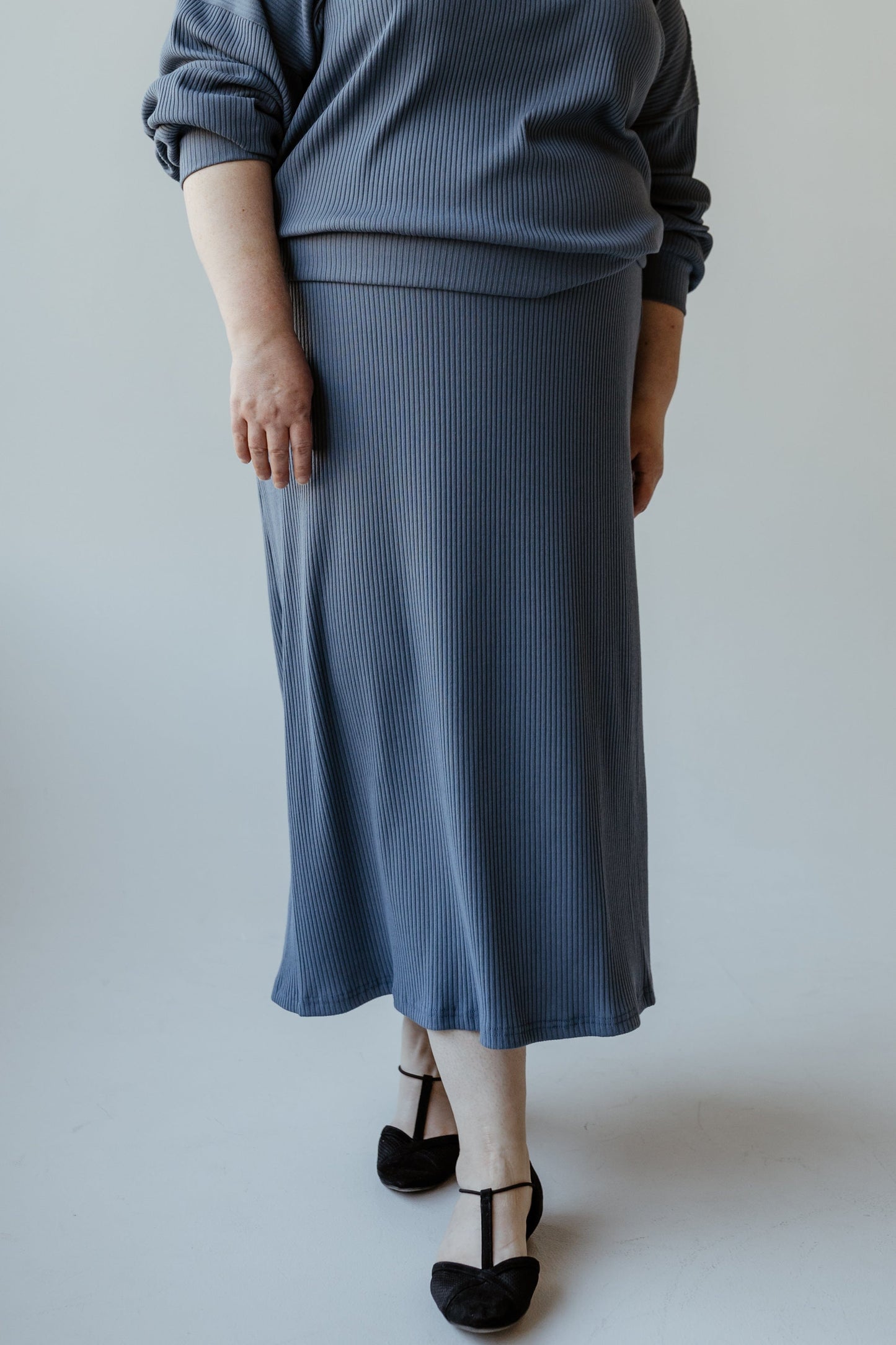 A person wearing the Ribbed Knit Midi Length Pencil Skirt in Adrift by Mikarose, paired with a ribbed knit blue sweater and black shoes, stands on a light background.
