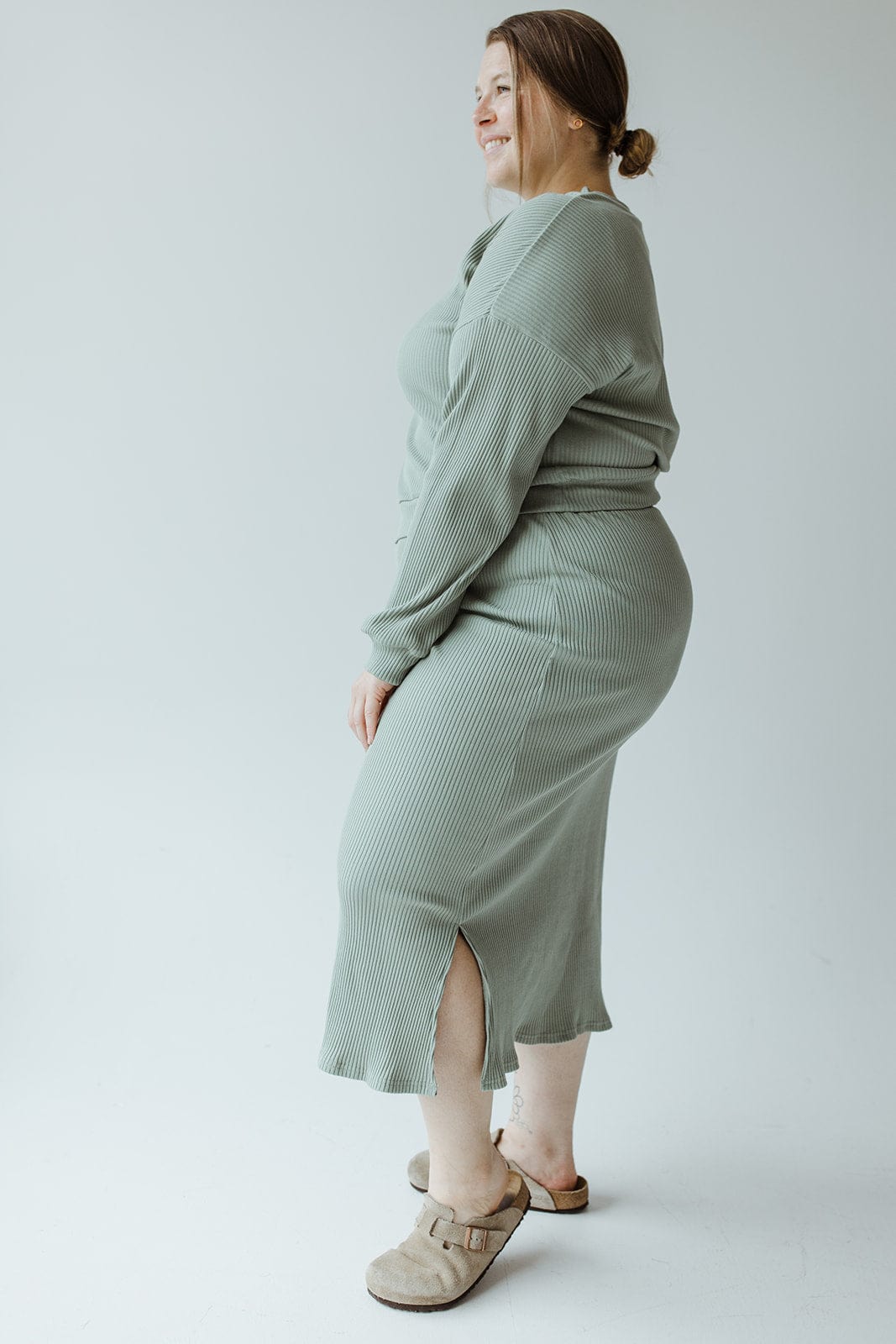 A woman in a Mikarose ribbed knit midi-length pencil skirt in Faded Jade and beige clogs stands sideways against a plain background, smiling.