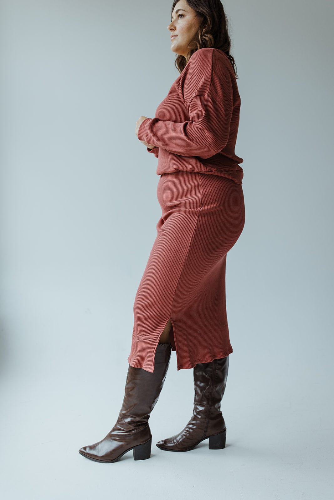 A person stands sideways wearing a rust-colored ribbed sweater and the Mikarose Ribbed Knit Midi Length Pencil Skirt in Smokey Rosewood, featuring slits, paired with brown knee-high boots, against a plain gray background.