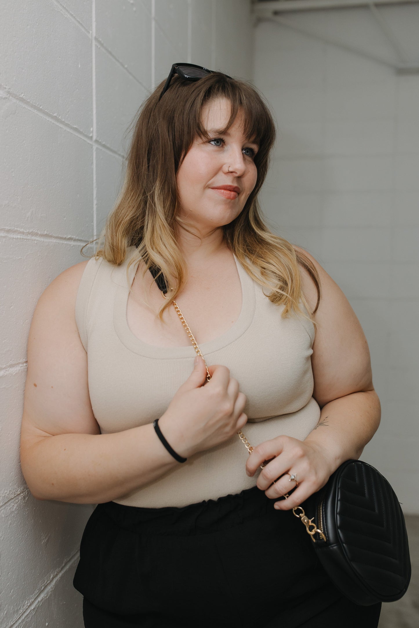 Woman holding the round quilted cross-body purse in black while she is wearing a beige top and black skirt, standing against a tiled wall.