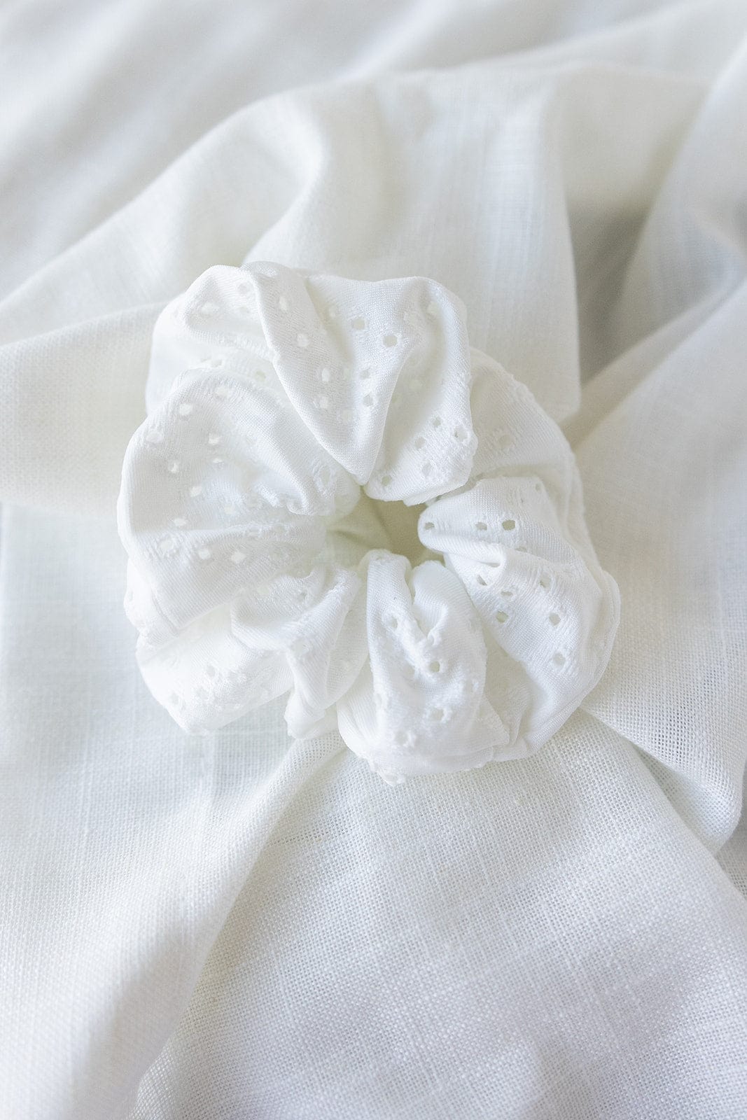 A white Eyelet Lace Scrunchie with delicate patterns is elegantly placed on a matching textured cloth background, perfect as a versatile hairstyle accessory.