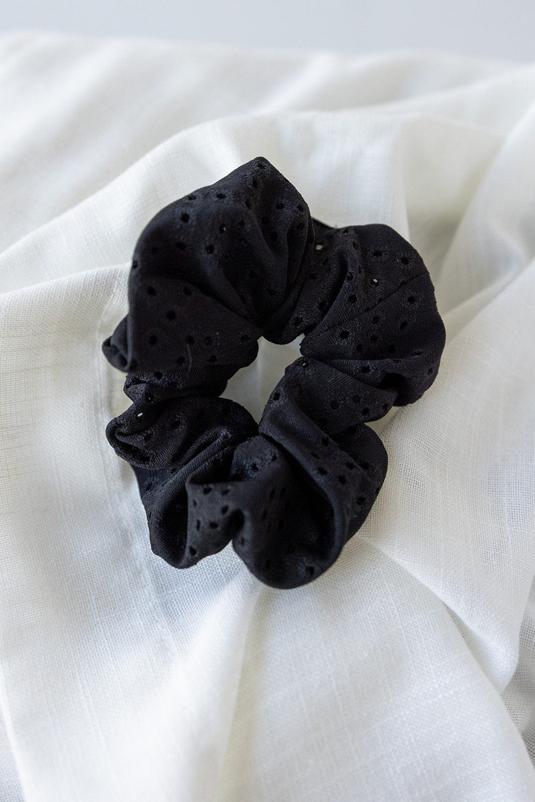 The black scrunchie, featuring a delicate eyelet lace design, elegantly rests on a white fabric background. This chic hair accessory is both stylish and versatile.