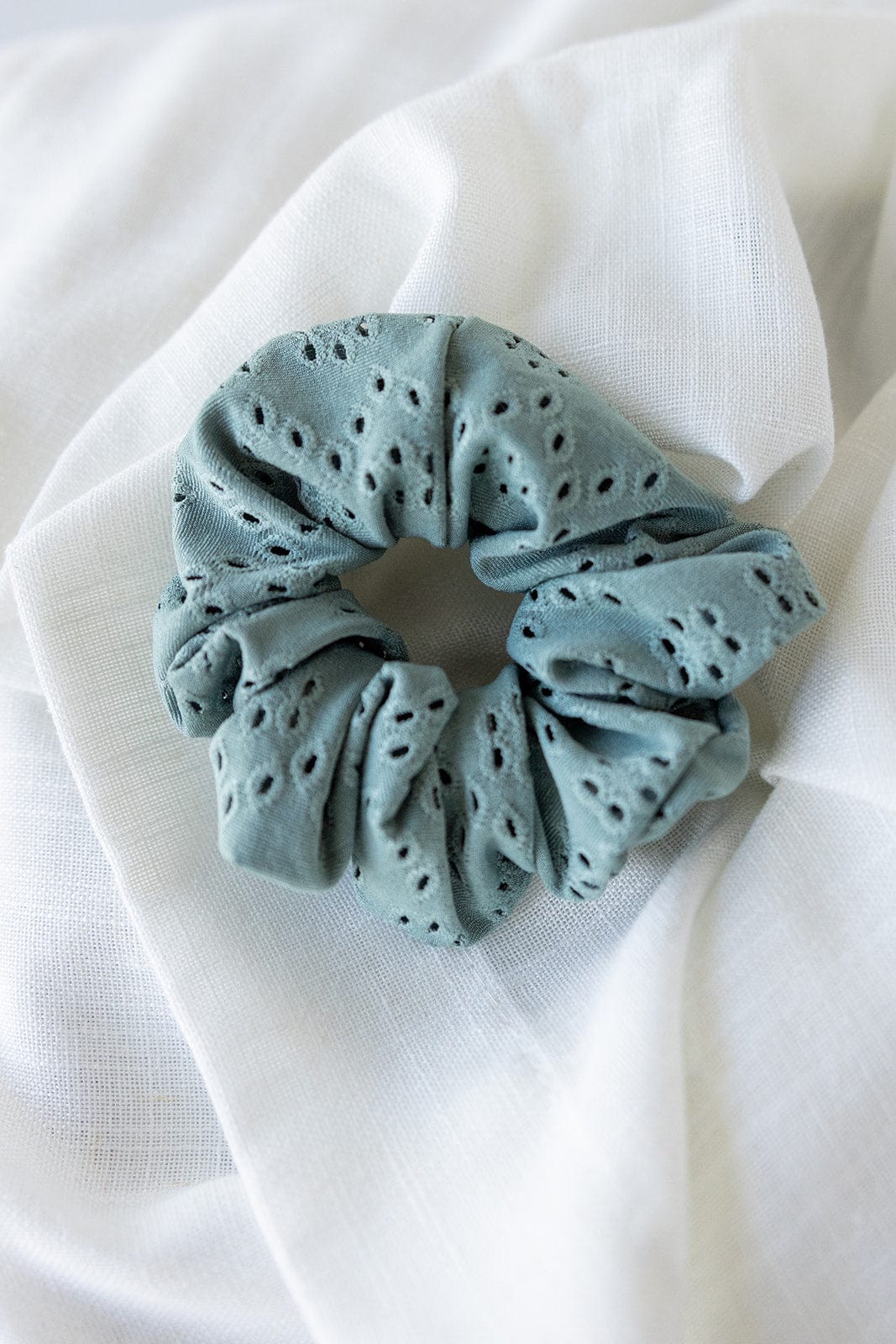 A chic ponytail is perfectly complemented by a sage green eyelet lace scrunchie resting on white fabric.