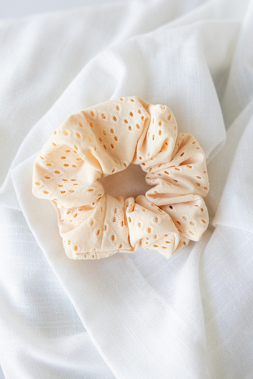 A stylish accessory, this peach-colored eyelet lace scrunchie gently rests on white fabric, adding a touch of elegance.
