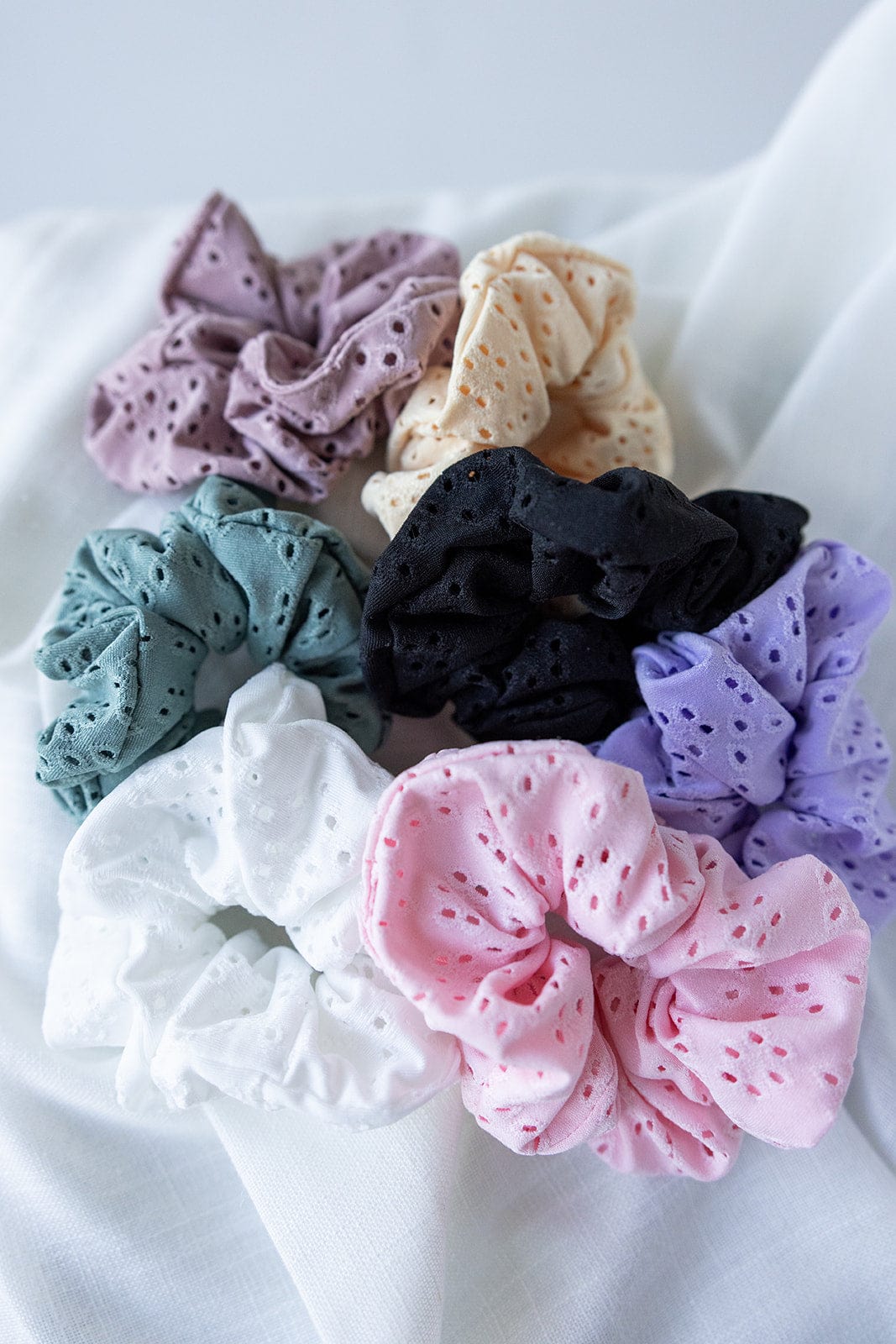 A collection of eight vibrant hair accessories with eyelet patterns, including a standout black scrunchie, elegantly arranged on a white fabric.