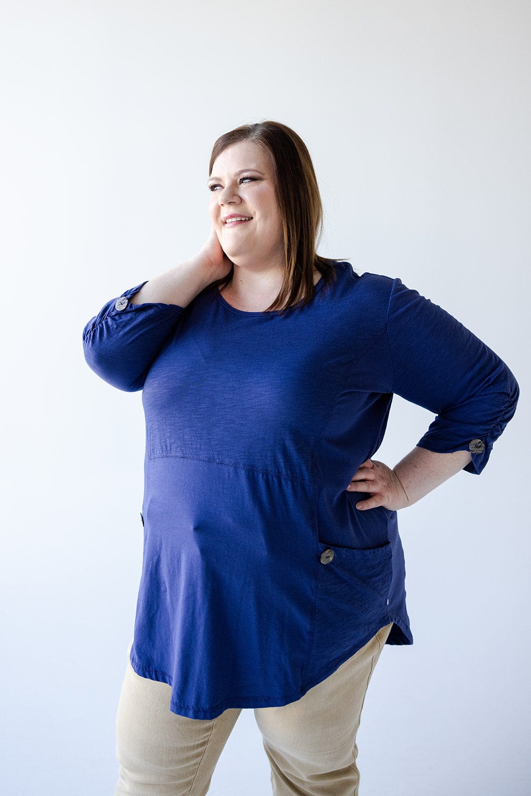 A woman wearing a Love Marlow 3/4 Ruched Sleeve Tunic with Pockets in Marine and beige pants stands while smiling, with one hand touching her neck and the other resting on her hip. The background is plain and white.