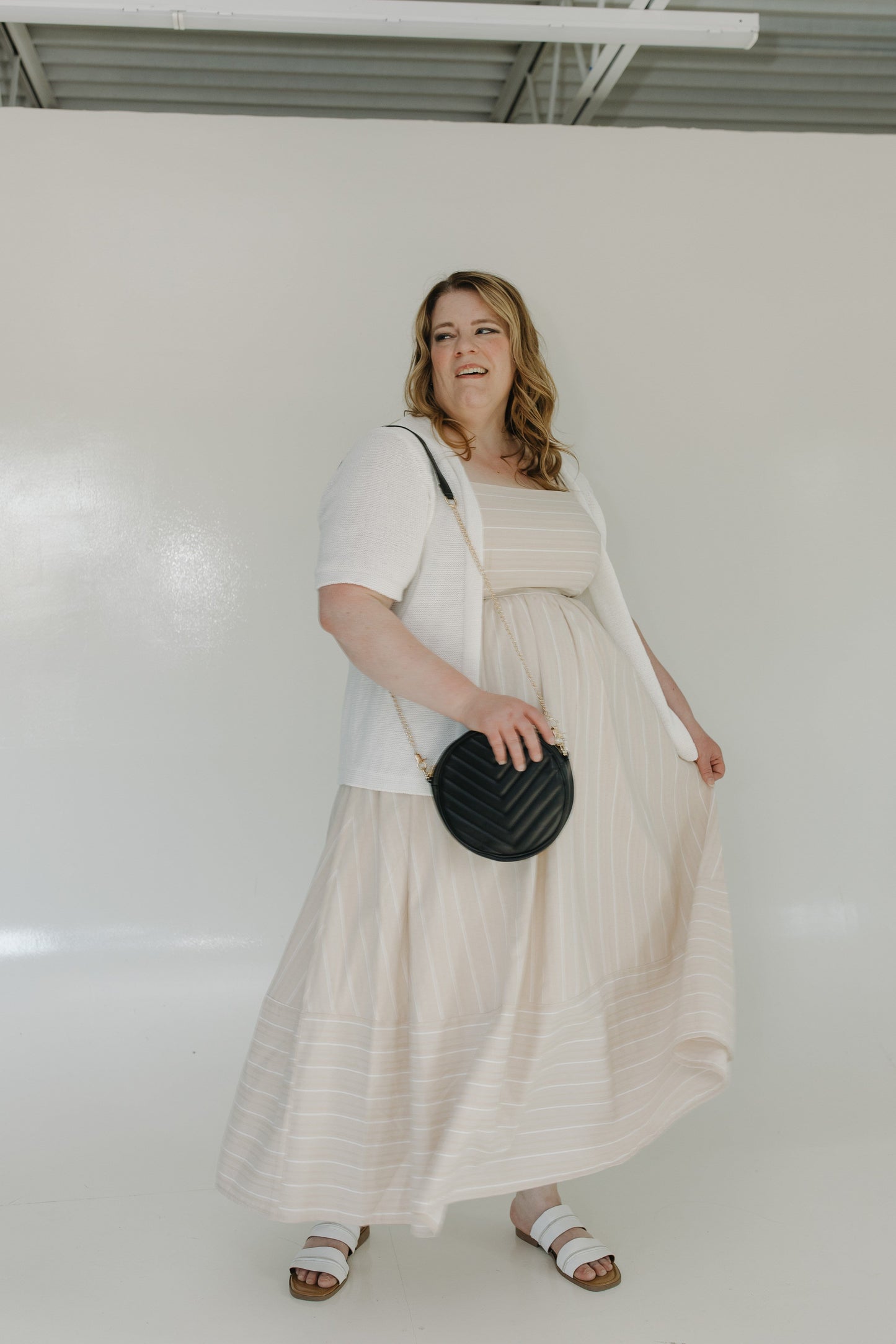 Woman holding the round quilted cross-body purse in black while she is wearing a beige top and black skirt, standing against a tiled wall.