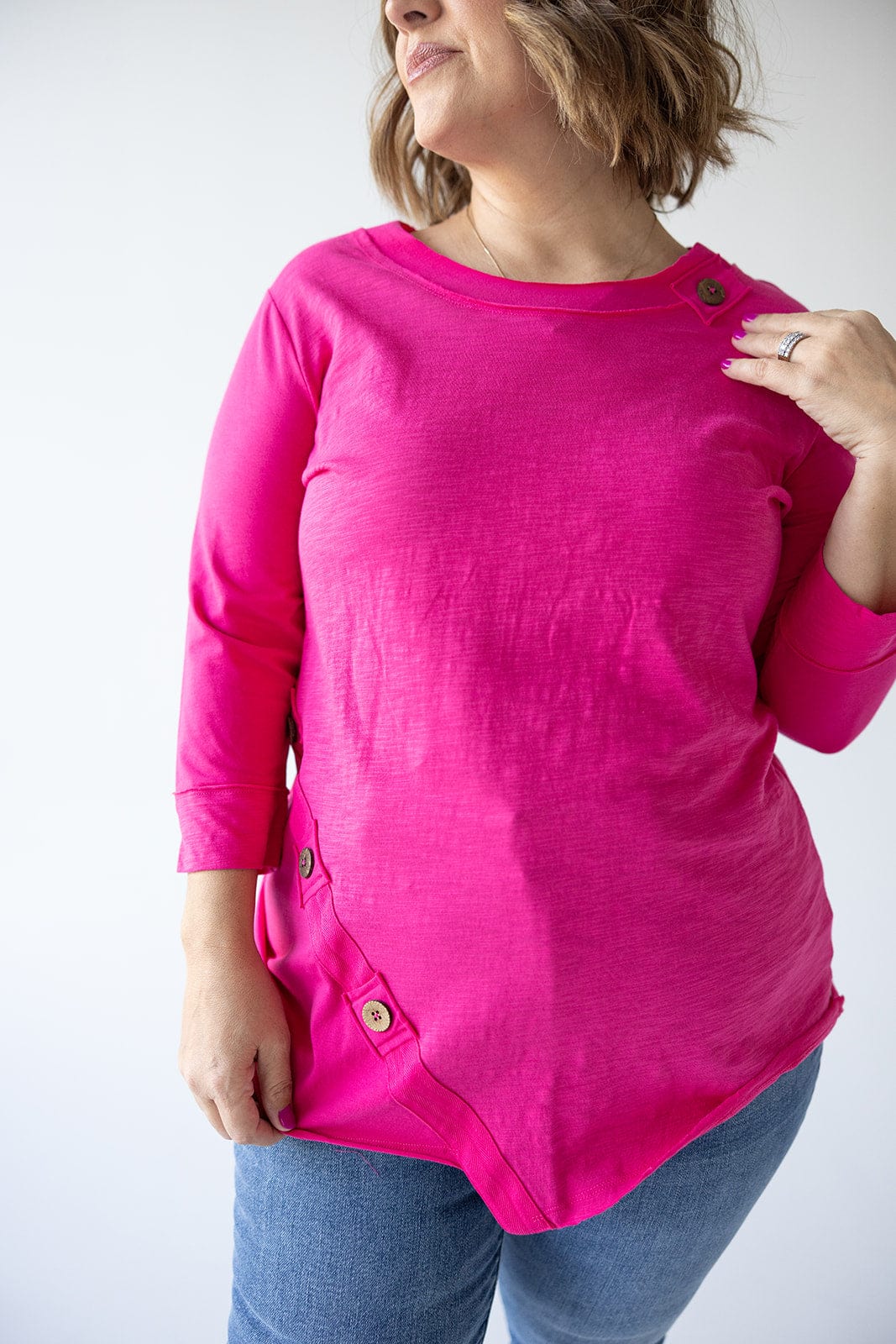 A woman wearing a bright pink, 3/4 SLEEVE TUNIC WITH BUTTON DETAIL IN GLAM PINK by Love Marlow, paired with blue jeans, poses with one hand near her shoulder and the other relaxed by her side.