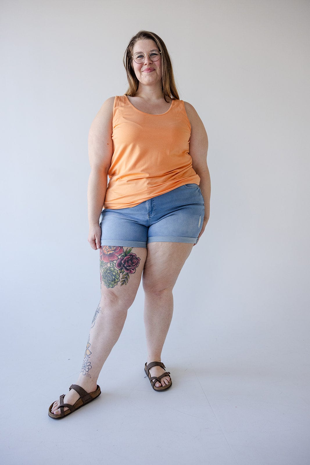 A person stands confidently in a peach tank top, Love Marlow SLIGHTLY DISTRESSED TUMMY CONTROL CUFFED SHORTS IN LIGHTWASH, and sandals. They have long hair, wear glasses, and have colorful floral tattoos on their right leg. The background is plain and light-colored.