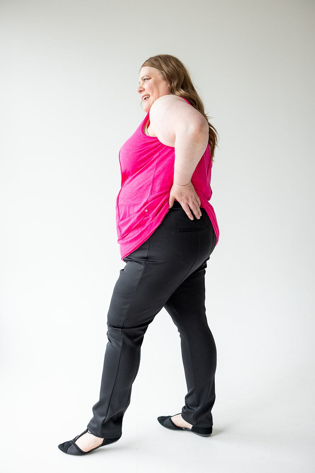 A woman in a pink top and black pants, with a slim straight leg design, stands sideways, smiling confidently with one hand on her hip in a bright room.
