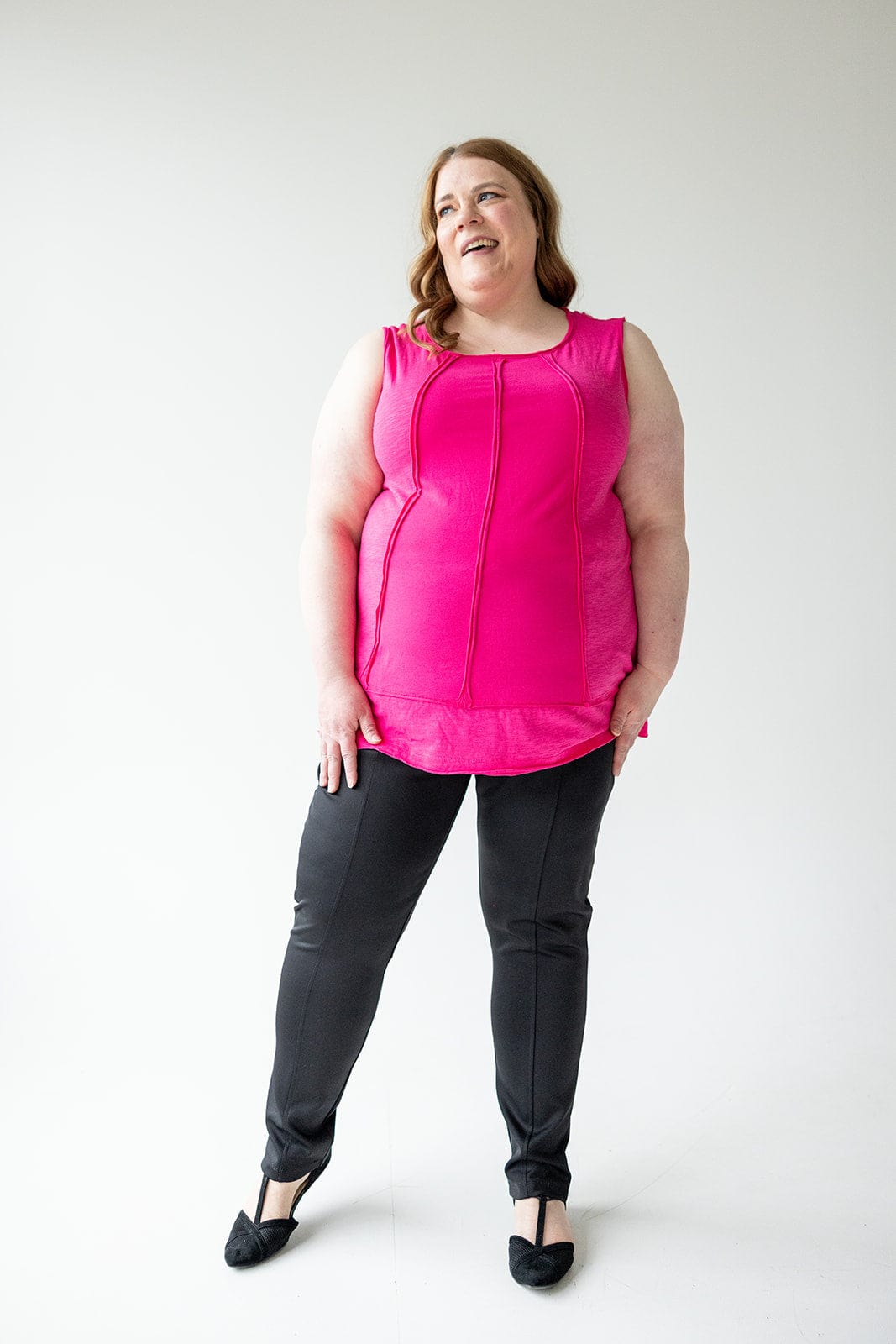 A woman in a pink sleeveless top and black pants with a slim straight leg stands against a plain background, smiling.