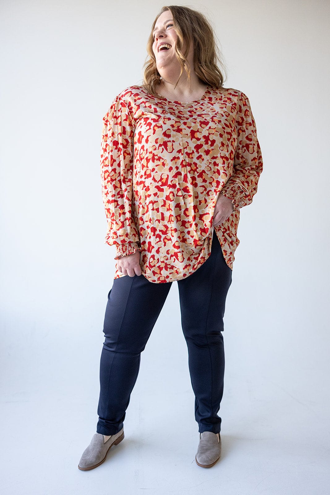 A person wearing a patterned blouse in sateen fabric, slim straight leg dark pants, and gray shoes stands smiling against a plain background.