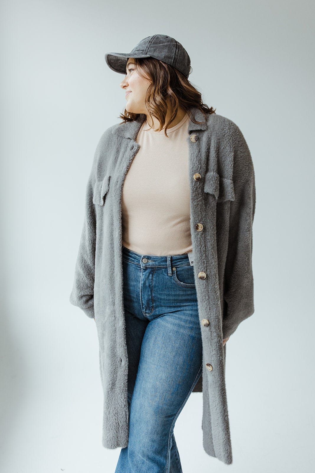 A woman stands against a plain background, wearing a steel grey shacket over a beige top, paired with jeans and a matching cap.