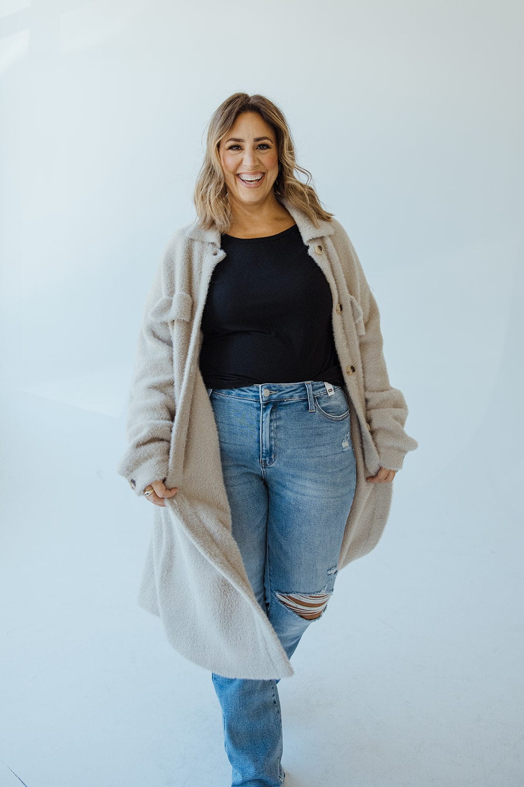 Person wearing a cozy shacket, black top, and ripped jeans stands in a light, minimalist setting.