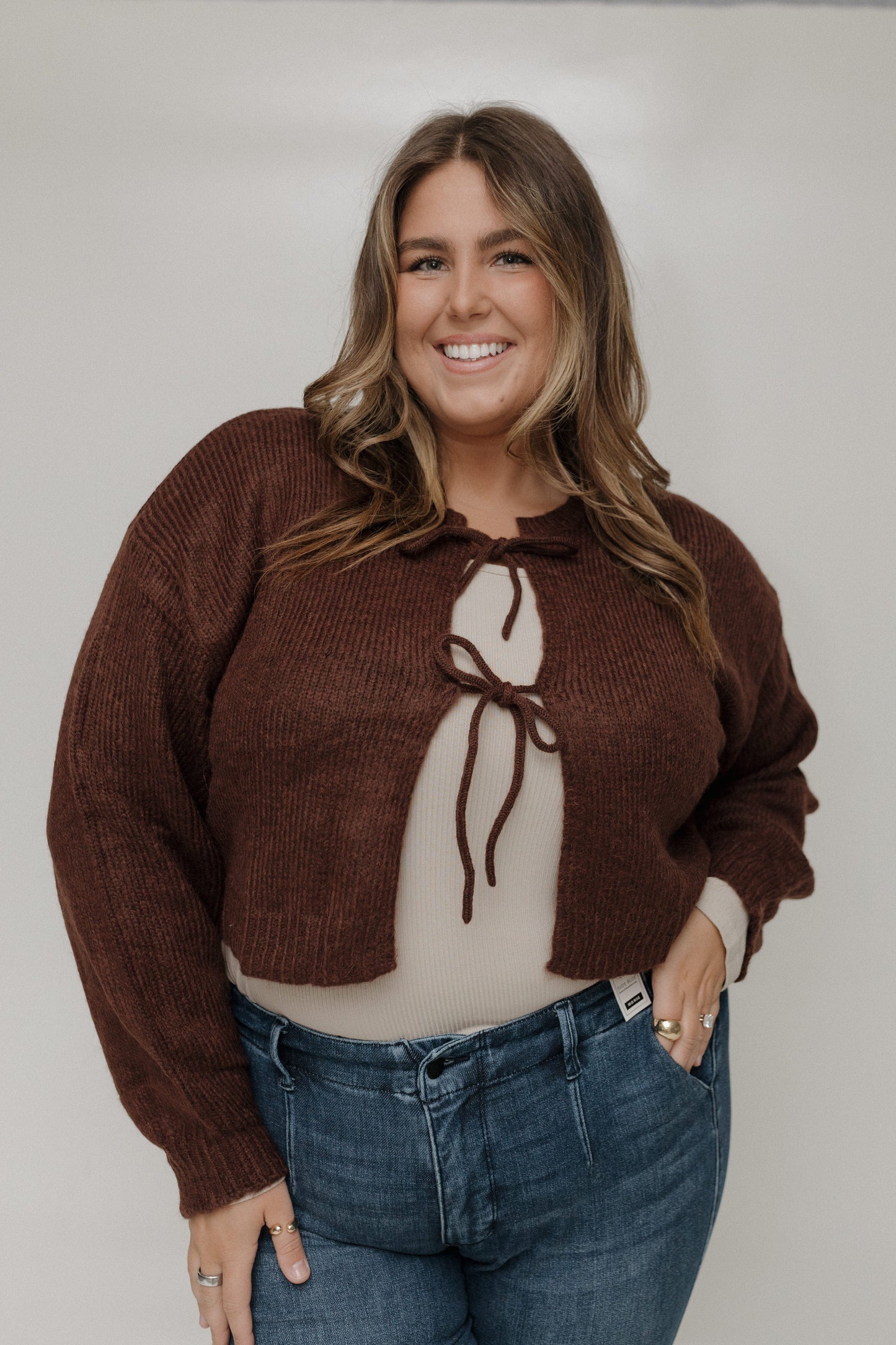 Woman wearing a brown sweater with a bow detail and blue jeans against a plain background