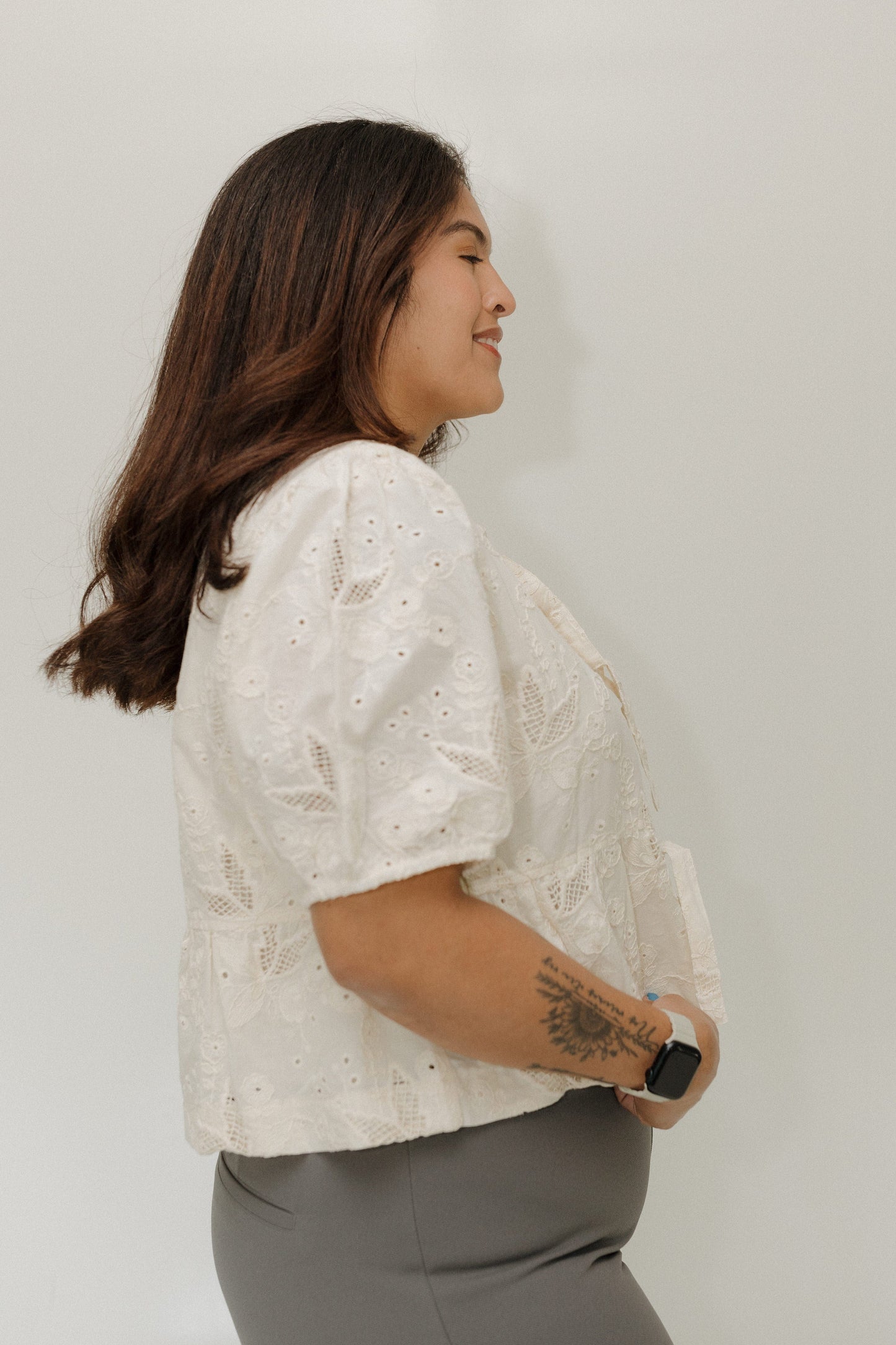 Woman wearing a white lace blouse and gray pants against a plain background