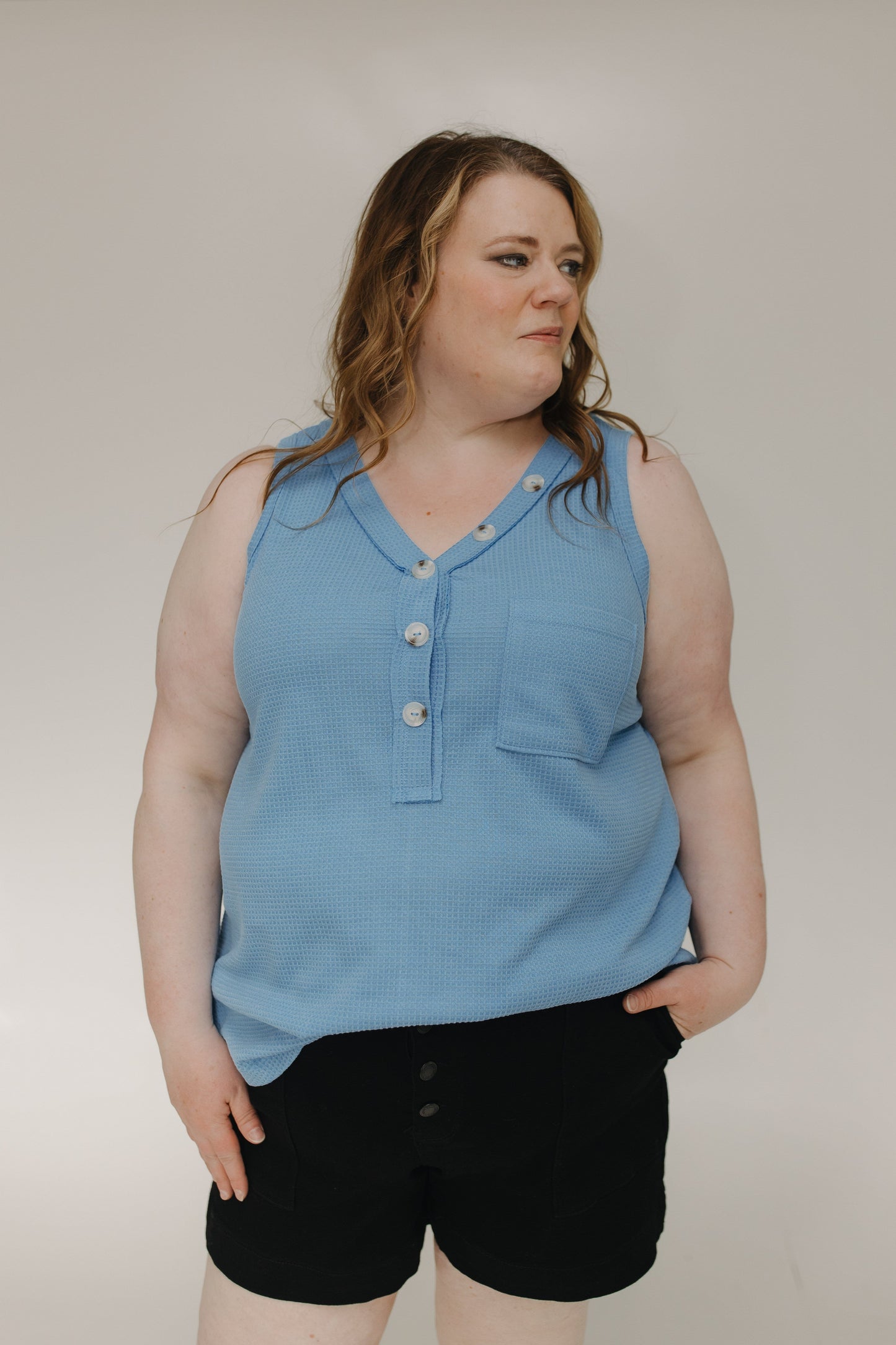Woman wearing a blue sleeveless shirt with buttons and black shorts against a plain background