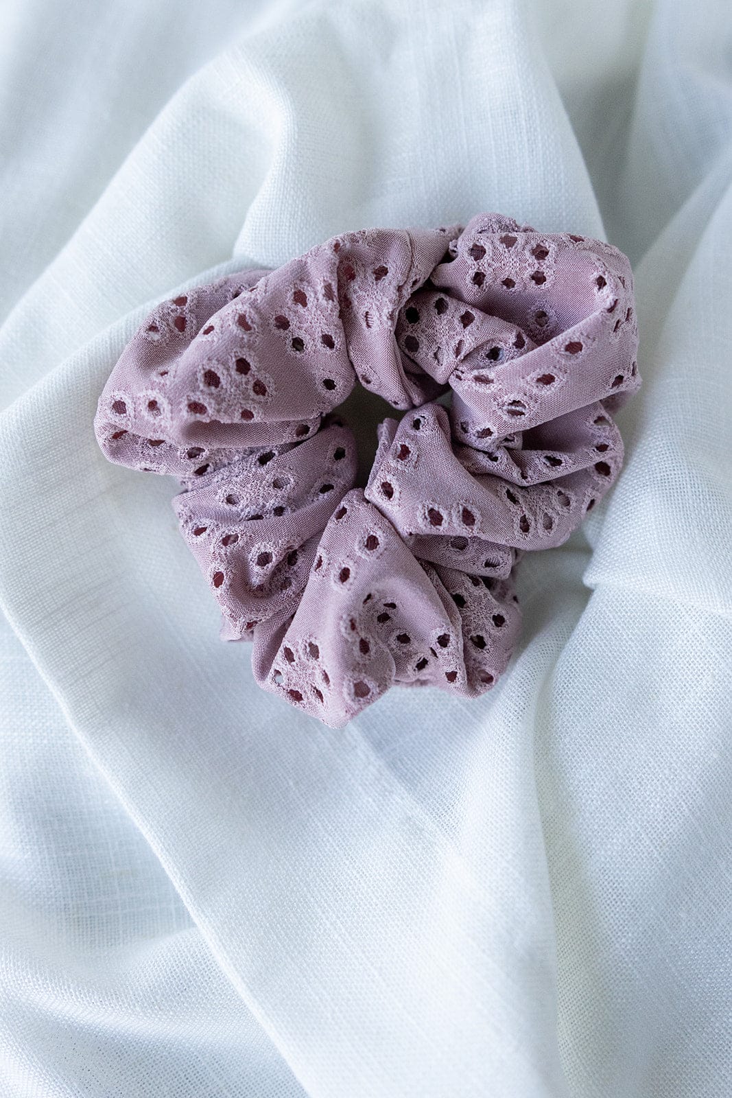 A mauve eyelet lace scrunchie delicately rests on a white fabric background.