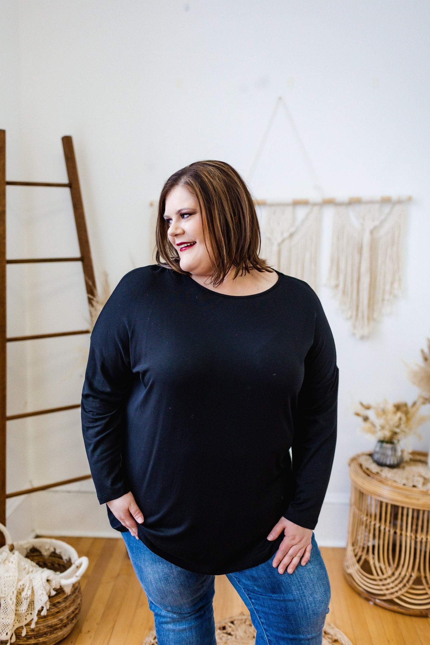 A person with shoulder-length hair wearing a Love Marlow BASIC DOLMAN TUNIC IN BLACK and blue jeans stands in a room with wooden and macrame decor.