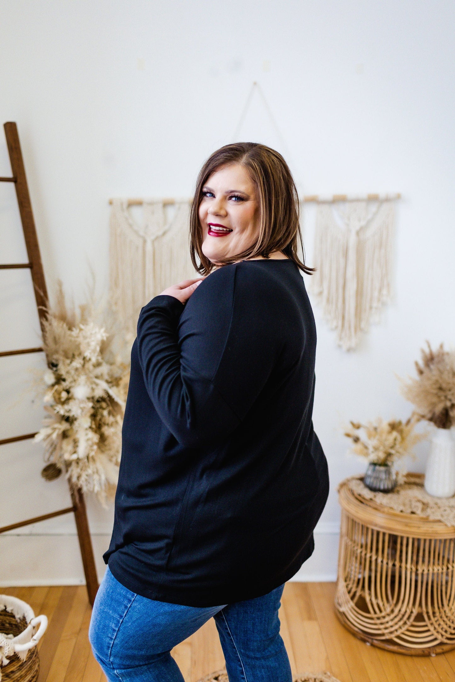 A person in a BASIC DOLMAN TUNIC IN BLACK from Love Marlow and blue jeans poses indoors, looking over their shoulder and smiling. The lightweight fabric of the tunic adds to their comfort. Dried floral arrangements and home decor items are visible in the background.