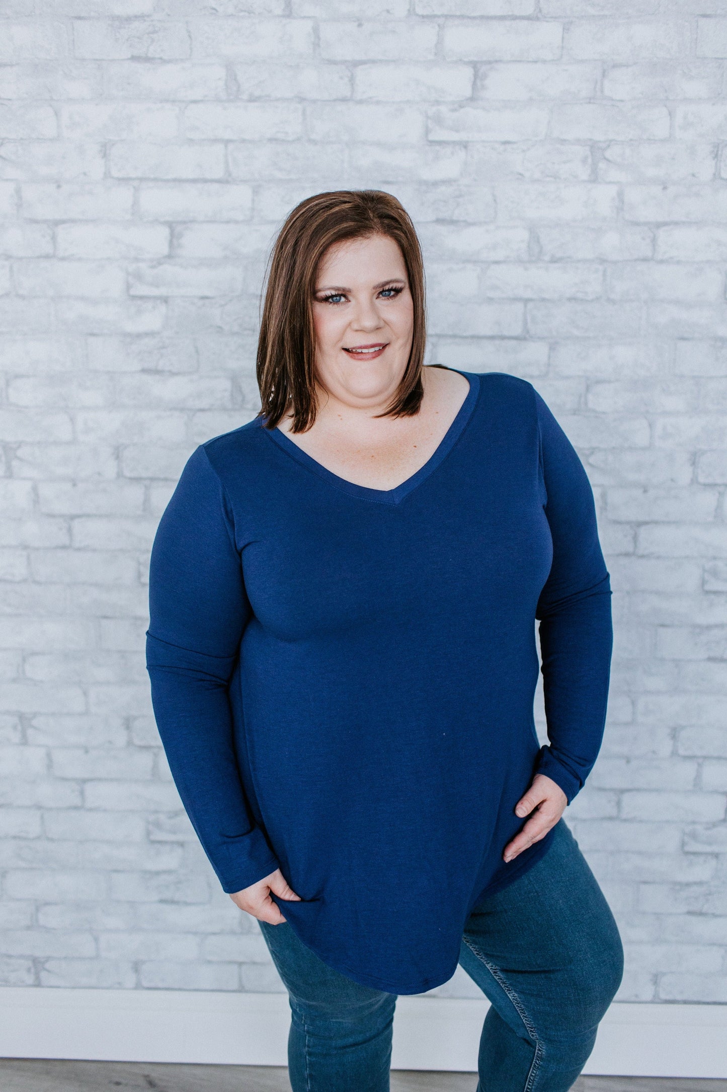 A person wearing a dark blue long-sleeve shirt and jeans stands in front of a white brick wall, facing the camera.