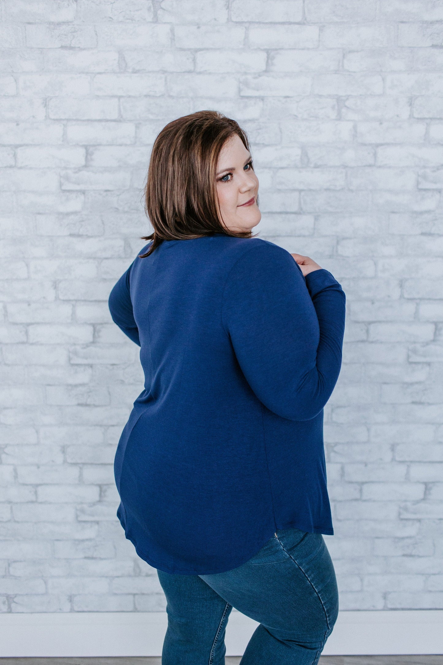 A person wearing a dark blue sweater and jeans stands in front of a white brick wall, looking over their shoulder.