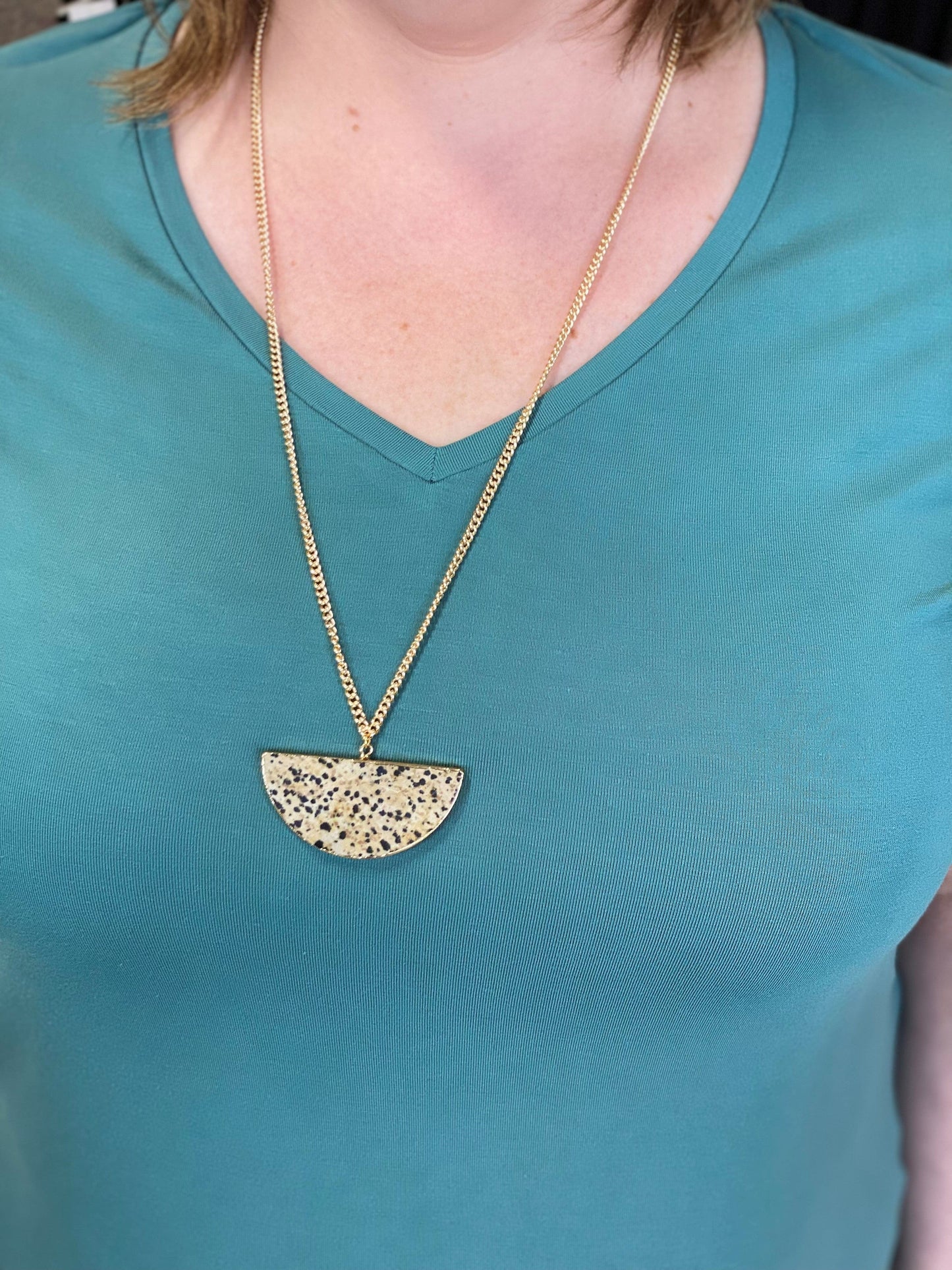 A person wearing a teal shirt and a long gold chain, reminiscent of Cleopatra's opulent style, flaunts a half-circle jasper pendant adorned with black speckles.