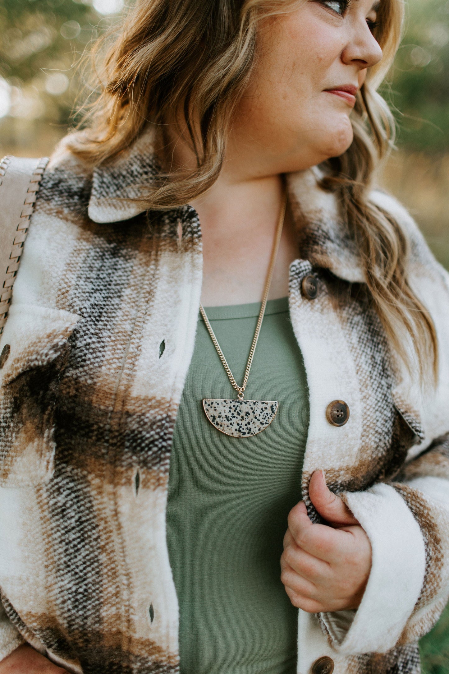 A woman in a plaid jacket over a green top is adorned with a half-moon pendant necklace, reminiscent of Cleopatra's elegance, as she gazes to the side.