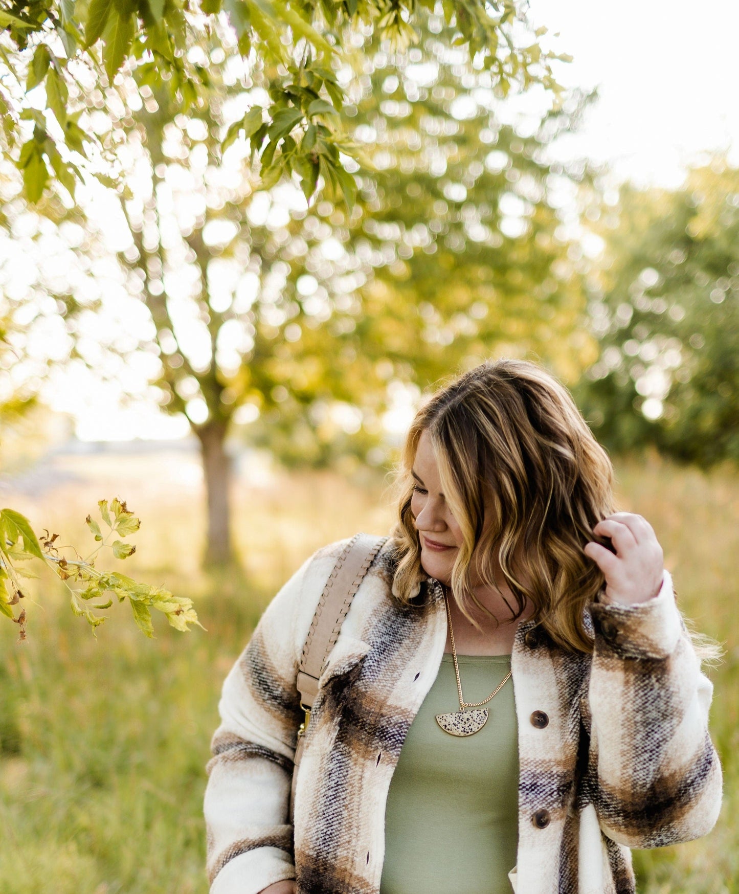 A woman with shoulder-length hair in a plaid jacket stands outdoors, brushing her hair back. A jasper pendant gleams against her outfit as she basks in the grassy area, with trees and sunlight filtering through.