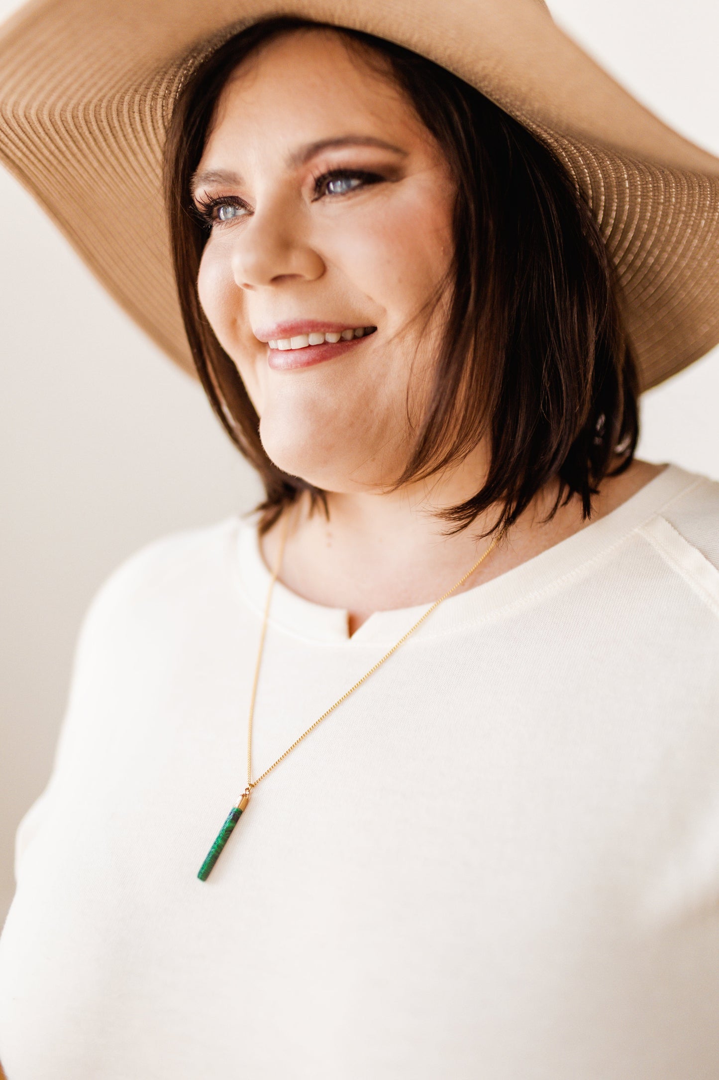 A woman, adorned with a wide-brimmed hat and a gold chain featuring a jasper pendant, smiles while gazing to the side.