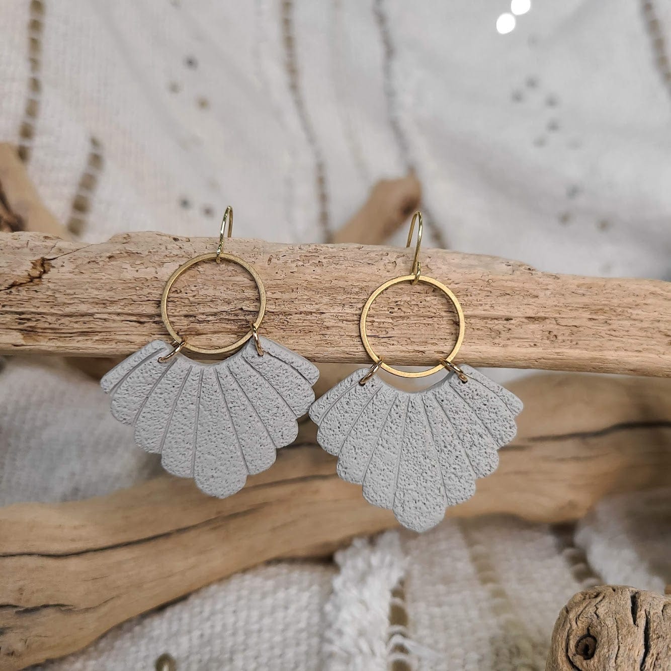 Gray shell-shaped earrings crafted from polymer clay with gold stainless steel hooks, elegantly hanging on driftwood against a textured fabric background.