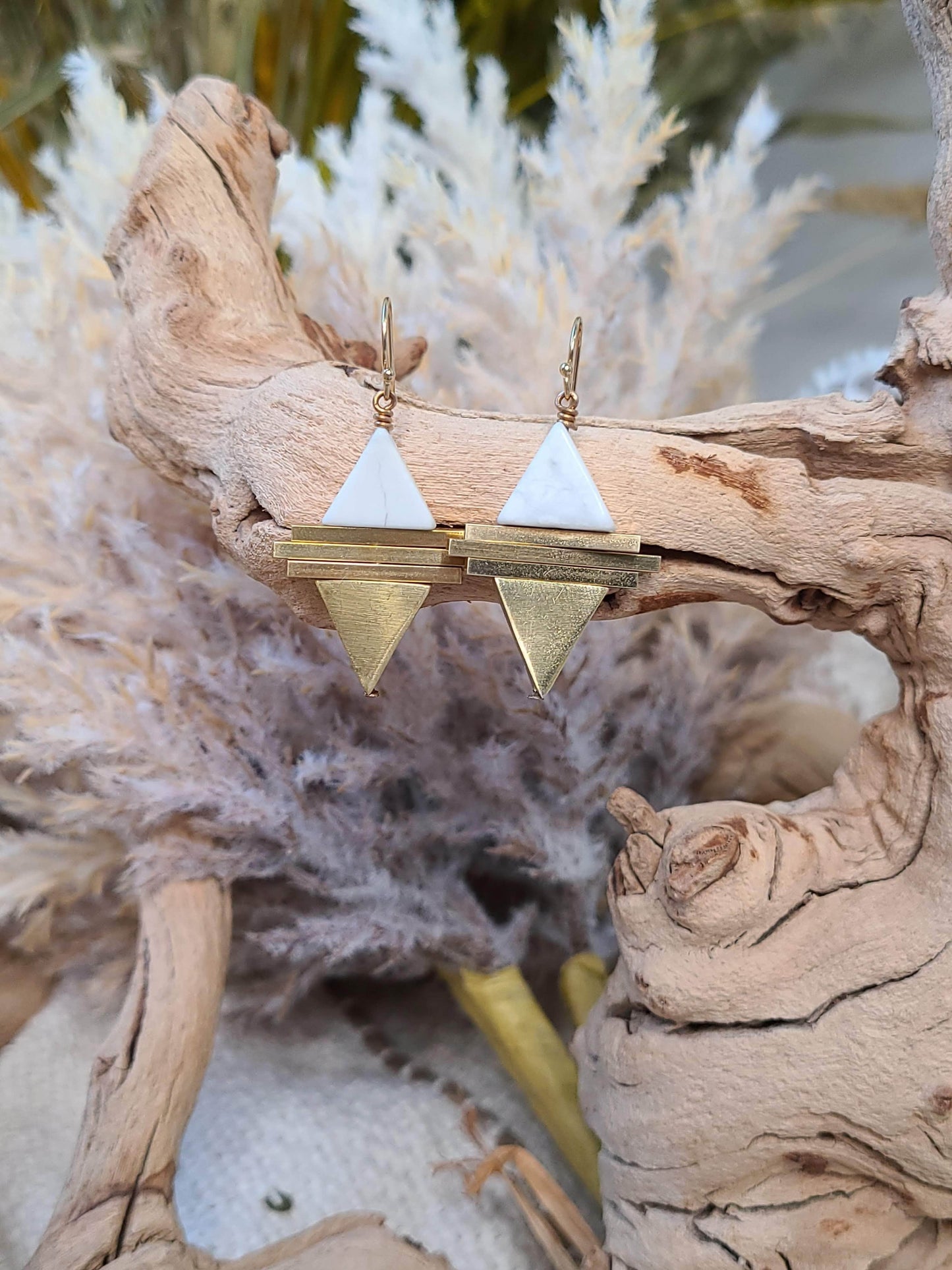 Gold and white geometric earrings with gold-filled ear wires are displayed on driftwood, featuring striking howlite triangles against a backdrop of pampas grass.