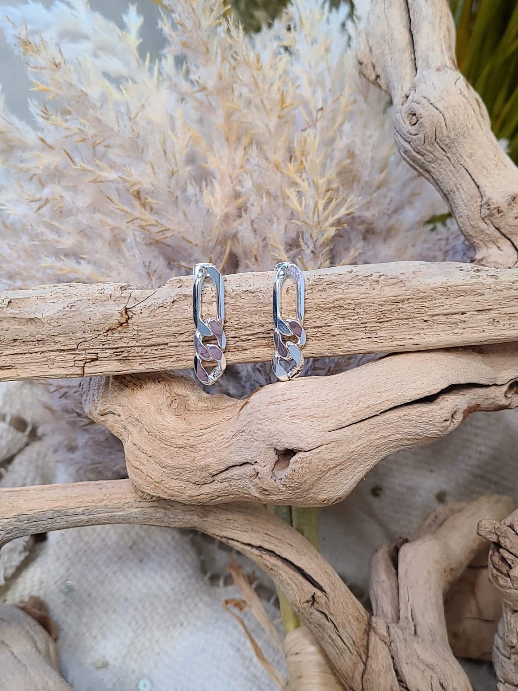 Silver-plated hoop earrings with a chain link design are artfully displayed on driftwood, accented by dried ornamental grass, epitomizing the essence of hypoallergenic jewelry.